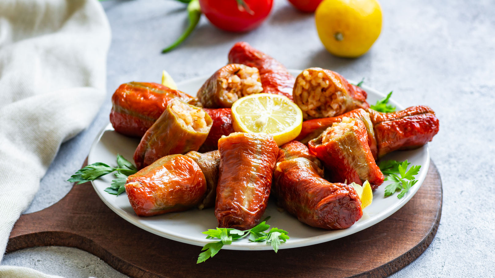Ein Teller mit gefüllten Paprika, die leicht knusprig und in verschiedenen Rottönen sind. Einige Paprika sind halb geöffnet, mit Sicht auf die Füllung. Eine Scheibe Zitrone und frische Kräuter dekorieren den Teller. Im Hintergrund sind frische Gemüselemente sichtbar.
