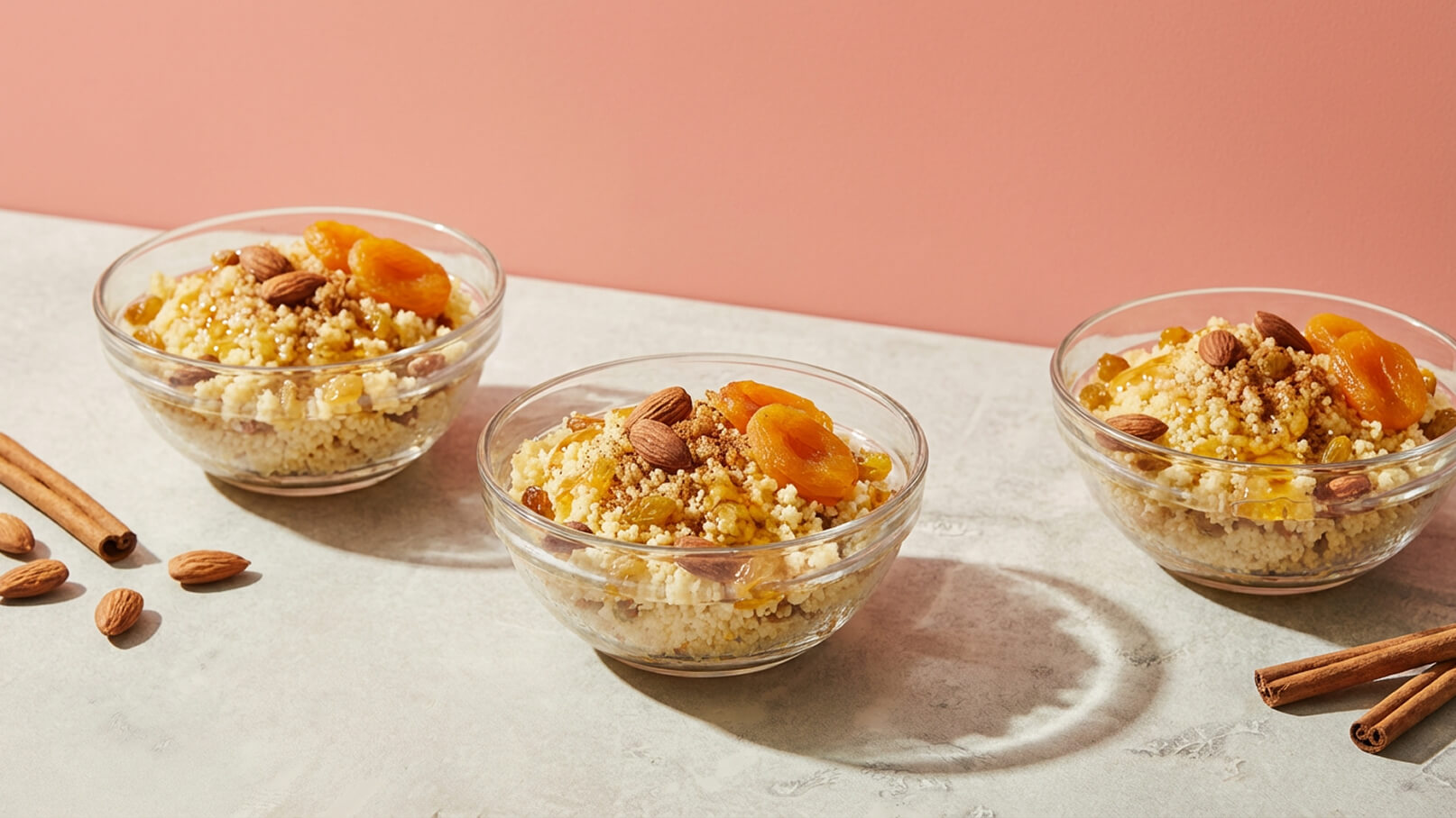 Drei gläserne Schalen mit Couscous, garniert mit getrockneten Aprikosen, Mandeln und Zimt. Der Hintergrund ist in einem sanften Rosa gehalten, und die Schalen stehen auf einer hellen Oberfläche.