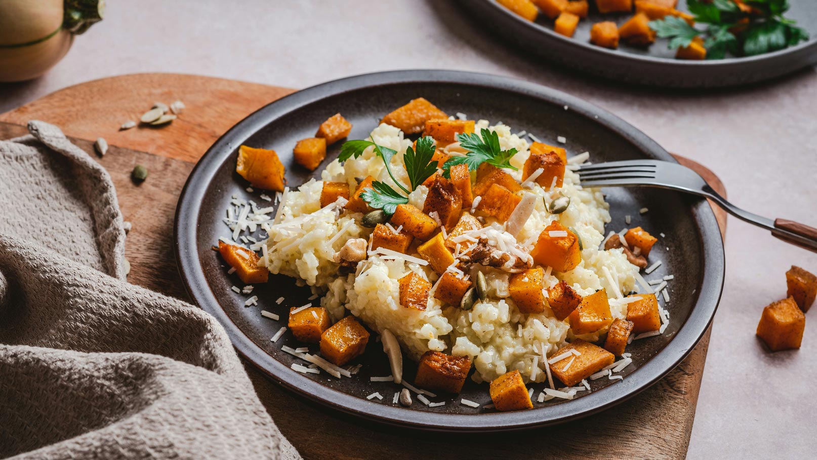 Ein dunkler Teller mit Risotto, garniert mit gebratenem Kürbis, Petersilie und geriebenem Käse. Der Kürbis ist goldbraun und in gleichmäßige Würfel geschnitten. Im Hintergrund liegt ein Holzbrett, auf dem eine Serviette und einige Kürbiskerne zu sehen sind.