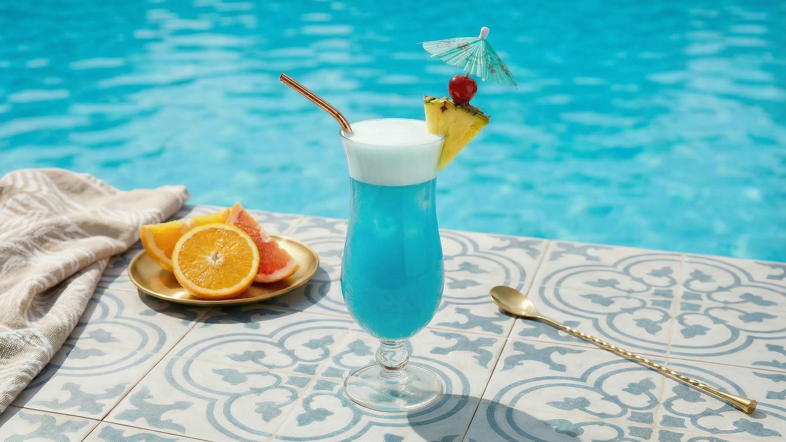 Ein blauer Cocktail in einem hohen Glas steht auf einem gefliesten Tisch. Der Drink ist mit einer kleinen, bunten Papierdeko, einer Ananas und einer Kirsche garniert. Neben dem Getränk liegen frische Früchte auf einem goldenen Teller. Im Hintergrund ist ein blauer Pool zu sehen.
