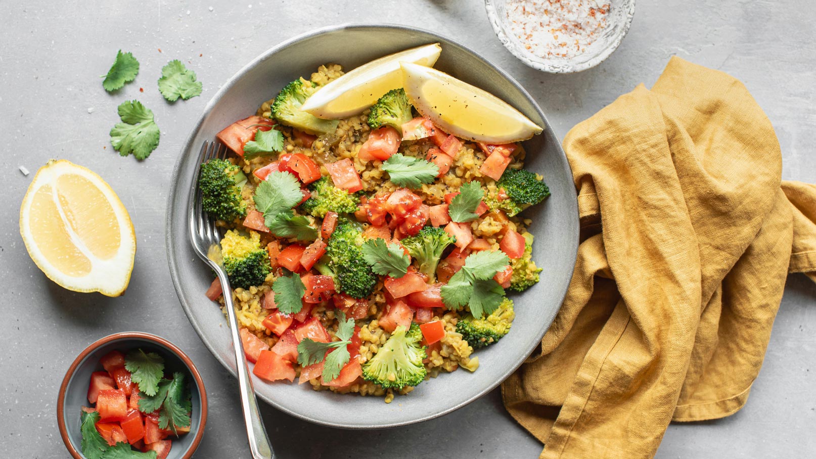 Eine Schüssel mit buntem Gemüsegericht aus Brokkoli, Tomaten und Koriander, garniert mit Zitronenscheiben. Neben der Schüssel befindet sich eine kleine Schale mit gehacktem Gemüse und Koriander sowie ein goldener Stoff.