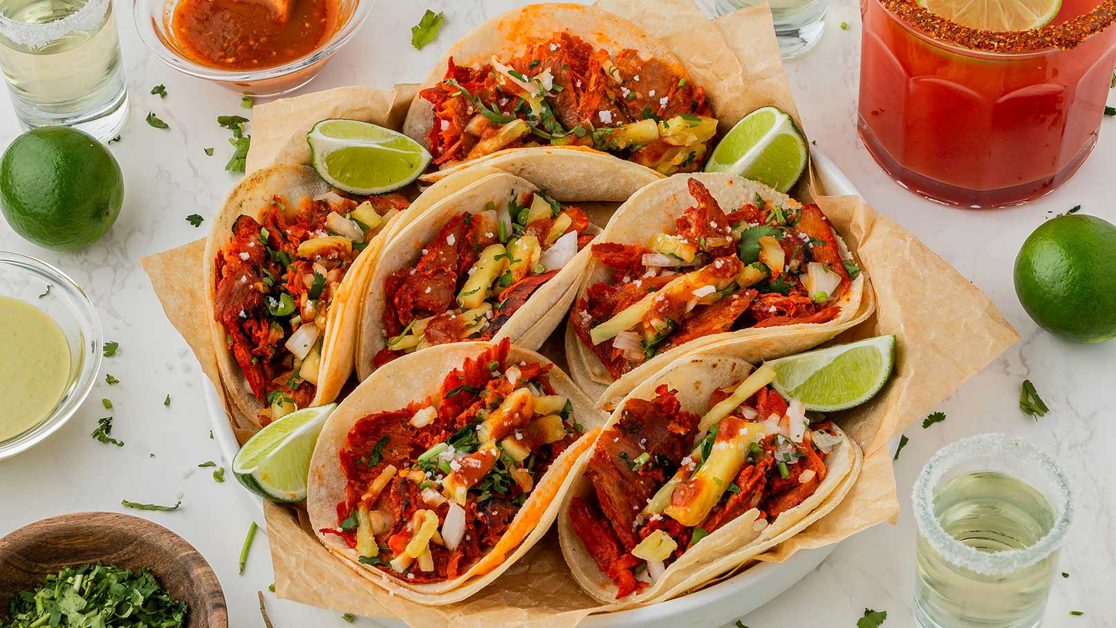 Auf einem Teller sind mehrere Tacos aufgereiht, gefüllt mit zarten, gewürzten Fleischstücken, frischem Gemüse und Kräutern. Limettenscheiben und kleine Schälchen mit Soßen sind ebenfalls angeordnet, die eine appetitliche Präsentation der Speise zeigen.