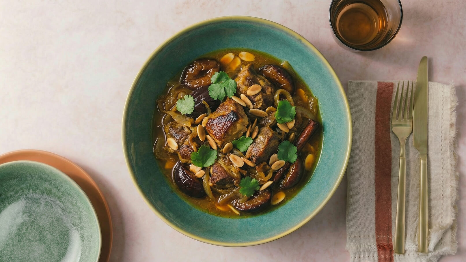 Ein tiefes grünes Geschirr enthält ein herzhaftes Gericht mit Fleischstücken, würzigen Zwiebeln und Datteln, garniert mit Koriander und gerösteten Mandeln. Daneben steht ein kleines Glas. Die Anrichtung ist ansprechend und einladend.