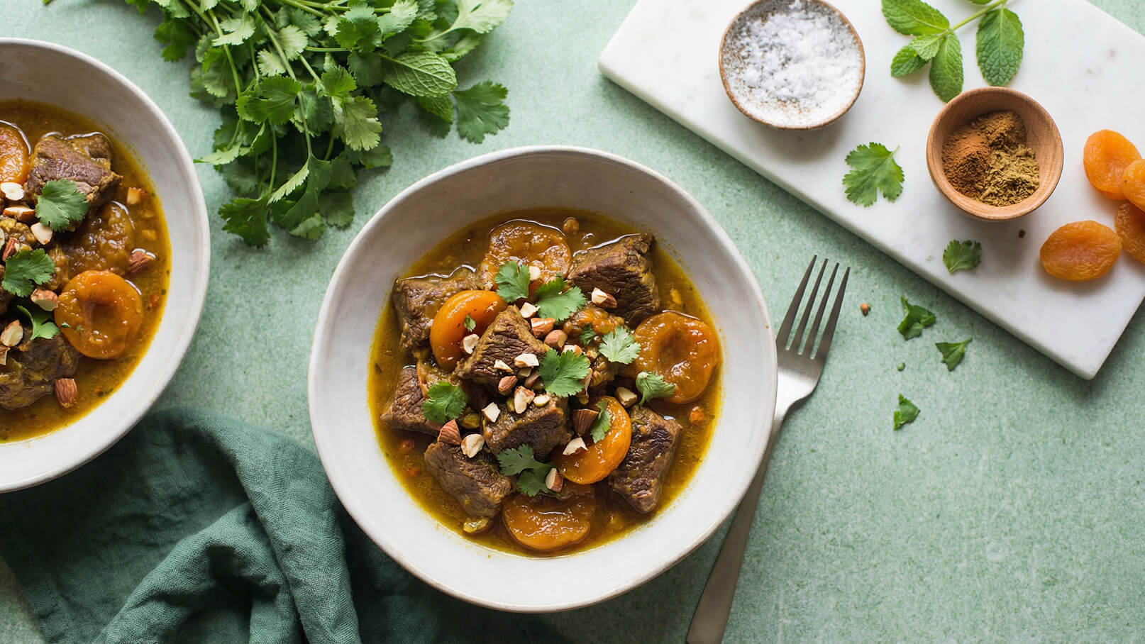 Eine Schüssel mit herzhaftem Eintopf aus zartem Fleisch, getrockneten Aprikosen und frischen Kräutern, garniert mit gehackten Nüssen. Daneben ein kleines Brett mit Gewürzen und getrockneten Aprikosen. Der Hintergrund ist in sanftem Grün gehalten.
