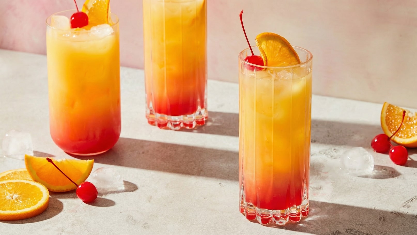 Drei hohe Gläser mit bunten, fruchtigen Getränken stehen auf einer Tischoberfläche. Die Getränke bestehen aus Gelb, Orange und Rot und sind mit Eiswürfeln, Orangenscheiben und einer Kirsche dekoriert. Einige Orangenscheiben und Kirschen liegen daneben.