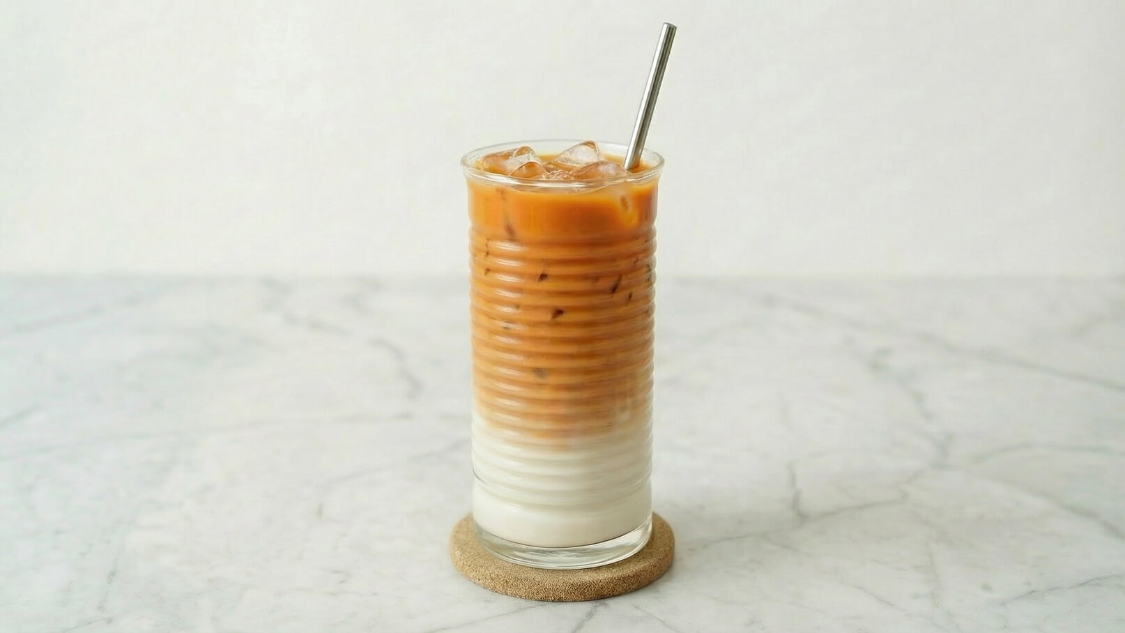 Ein Glas mit einem kalten Kaffeegetränk, das über Eiswürfeln serviert wird. Der Kaffee ist oben und die Milch darunter sichtbar, was einen schönen Farbverlauf bildet. Das Glas steht auf einem Untersetzer auf einer marmorierten Oberfläche.