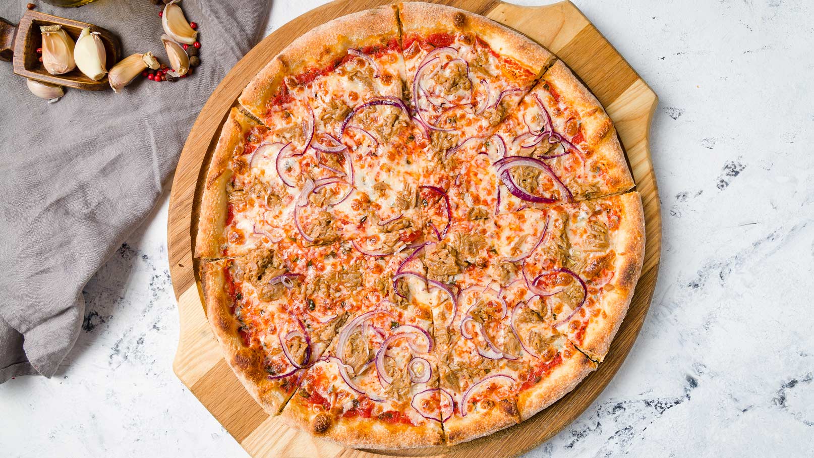 Eine frisch gebackene Pizza liegt auf einem Holzbrett. Sie ist mit Käse, roten Zwiebeln und Fleisch belegt und hat einen goldbraunen Rand. Die Pizza ist in Stücke geschnitten und befindet sich auf einem hellen Tisch.