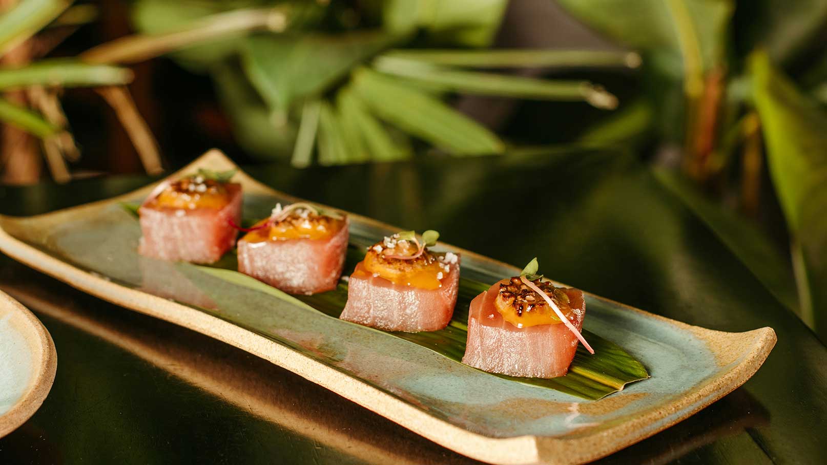 Auf einem stilvollen Teller liegen vier kleine Stücke Sushi. Jedes Stück besteht aus frischem Fisch mit einer köstlichen Topping-Garnierung, das auf einem Blatt platziert ist. Die Farben und Texturen des Sushis wirken appetitlich und ansprechend.