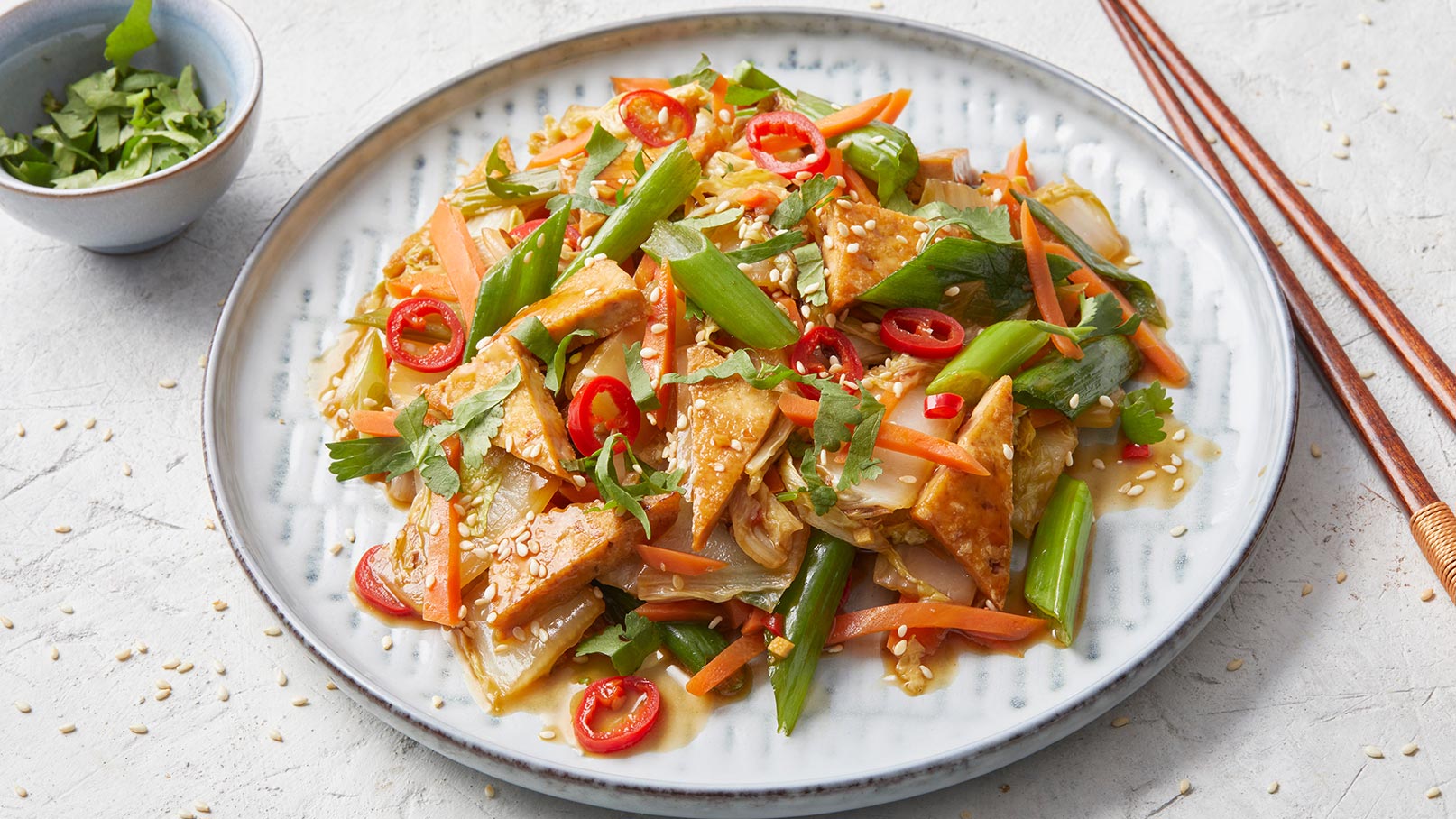 Ein Teller mit buntem Gemüsegericht, bestehend aus gebratenem Tofu, Karotten, Frühlingszwiebeln und Paprika, garniert mit Koriander und Sesam. Es sind auch rote Chili-Scheiben sichtbar. Im Hintergrund befindet sich eine kleine Schüssel mit frischen Kräutern.