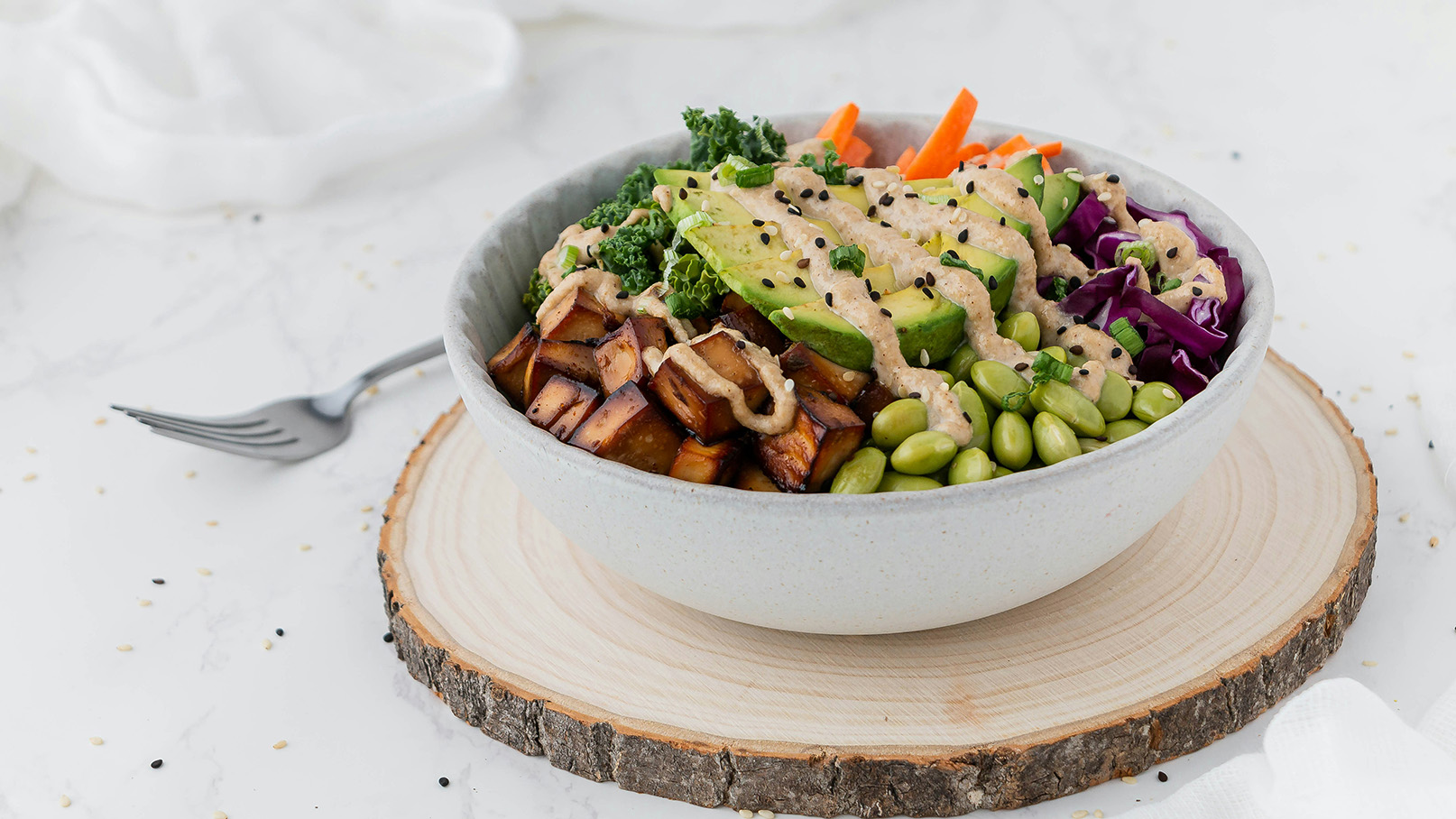 Eine Schüssel gefüllt mit einer bunten Gemüseauswahl, darunter Avocado, Karotten, Grünkohl und lila Kohl. Darauf befindet sich ein cremiges Dressing und geröstete Sesamsamen. Die Schüssel steht auf einem Holzuntersetzer.