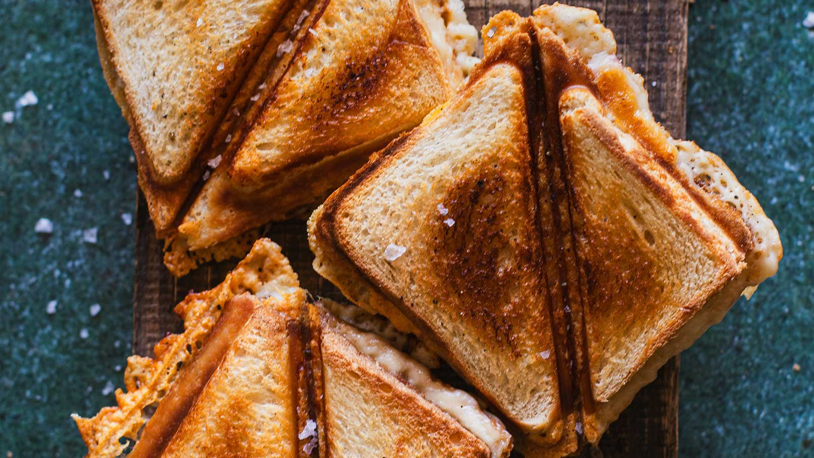 Ein Stapel goldbraun gerösteter Sandwiches auf einem Holzbrett. Die Sandwiches sind diagonal geschnitten und enthalten geschmolzenen Käse, der an den Seiten herausquillt. Einige Körner Salz sind auf der Oberfläche sichtbar.