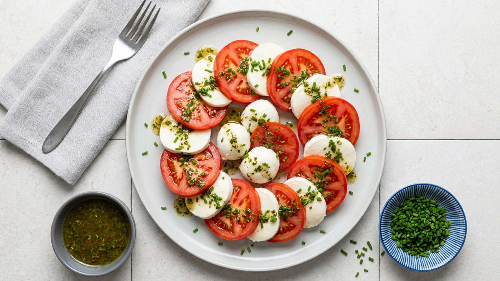 Ein runder Teller mit frischen Tomatenscheiben und Mozzarella-Kugeln, dekoriert mit Schnittlauch. Das Gericht ist durch ein grünes Kräuterdressing umgeben und präsentiert eine appetitliche Anordnung der Zutaten. Ein Besteck und ein Tuch liegen daneben.
