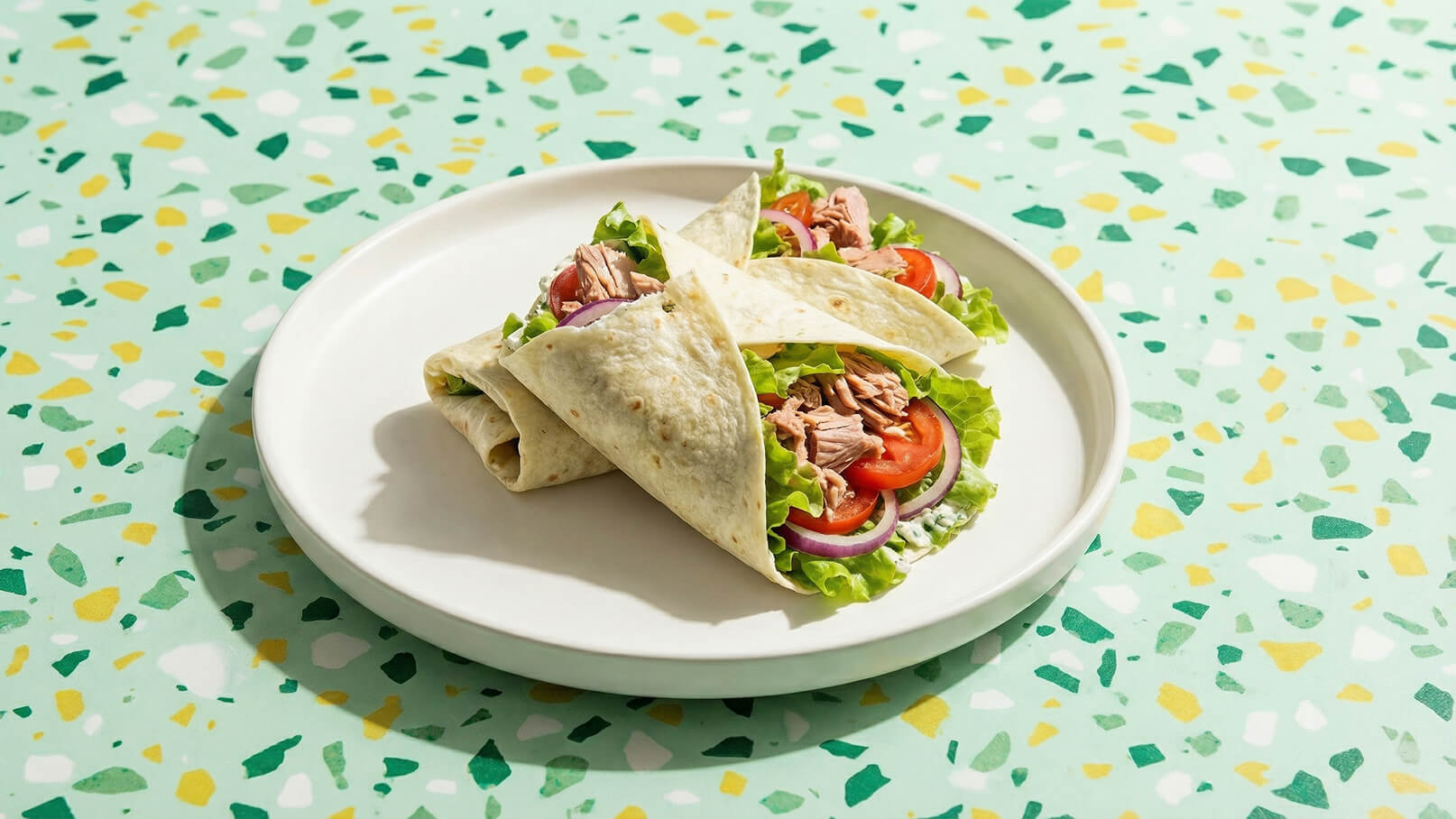 Auf einem weißen Teller liegen drei gefüllte Wraps. Sie bestehen aus Tortilla, Salat, Tomaten, roter Zwiebel und Thunfisch. Der Hintergrund ist in sanften Grüntönen mit gelben und weißen Akzenten gestaltet.