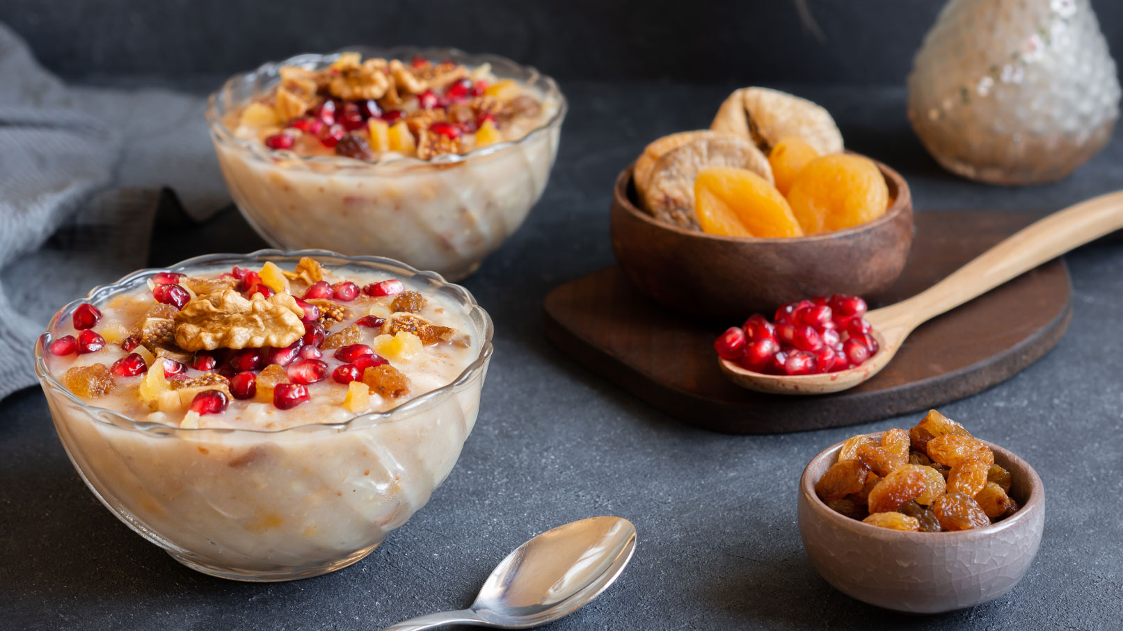 Zwei Schalen mit cremigem Pudding sind mit Granatapfelkernen, Walnüssen und Trockenfrüchten dekoriert. Daneben befinden sich eine Holzschale mit getrockneten Aprikosen und eine kleine Schüssel mit Rosinen. Im Hintergrund ist ein Löffel mit weiteren Granatapfelkernen zu sehen.