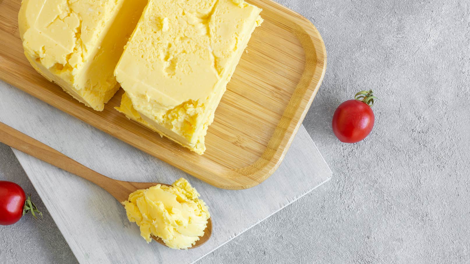 Ein Holzbrett zeigt einen Block Butter, von dem ein Stück abgeschnitten ist. Neben der Butter liegt ein Löffel mit Butterstückchen. Im Vordergrund ist eine kleine rote Tomate zu sehen. Der Hintergrund ist neutral und leicht strukturiert.