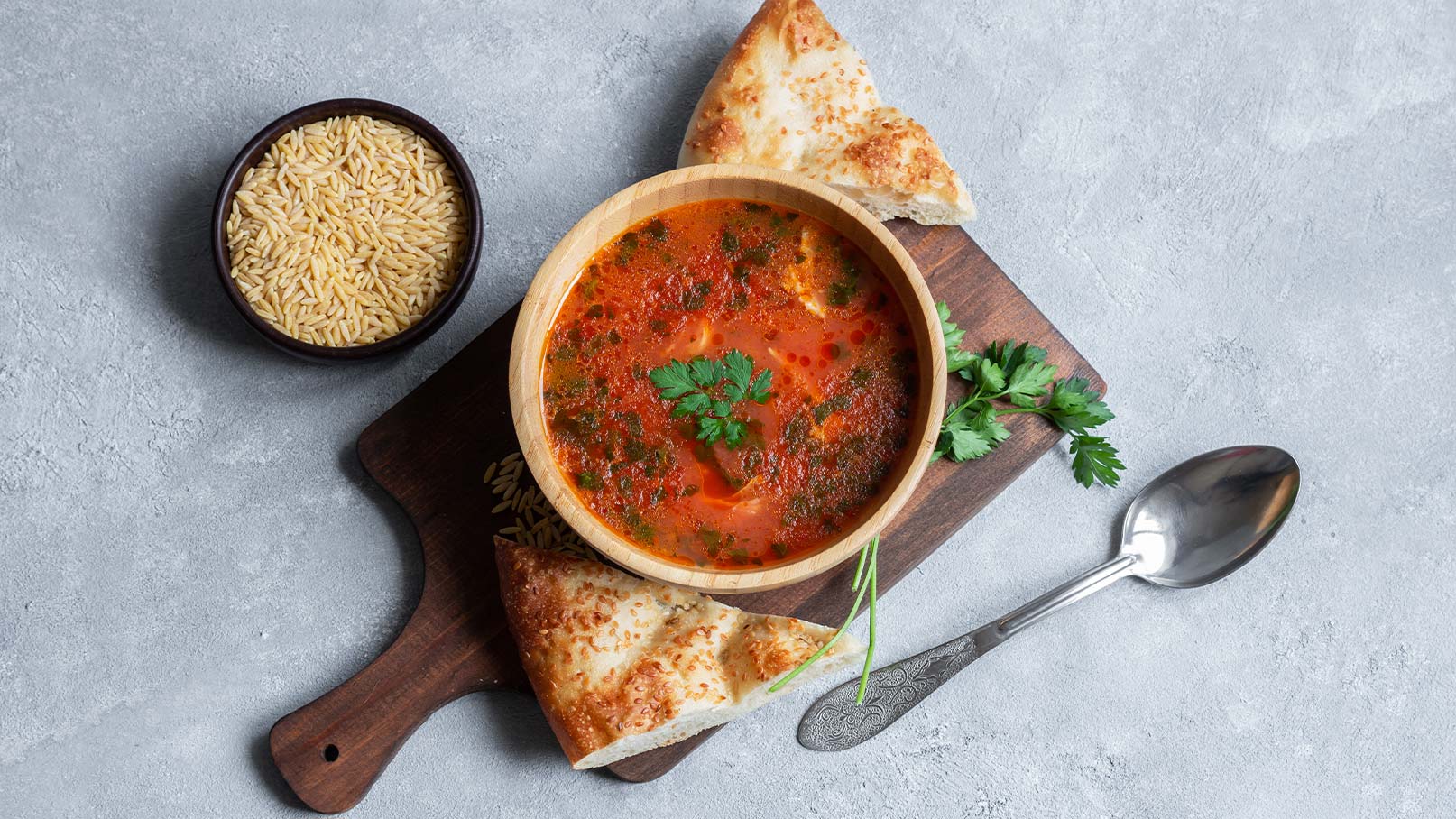 Eine Holzschüssel mit roter Suppe, garniert mit frischem Koriander, steht auf einem Holzbrett. Neben der Suppe liegen Stücke von knusprigem Brot und ein kleiner Schalen gefüllt mit ungekochten Reis. Der Hintergrund ist hell und schlicht.