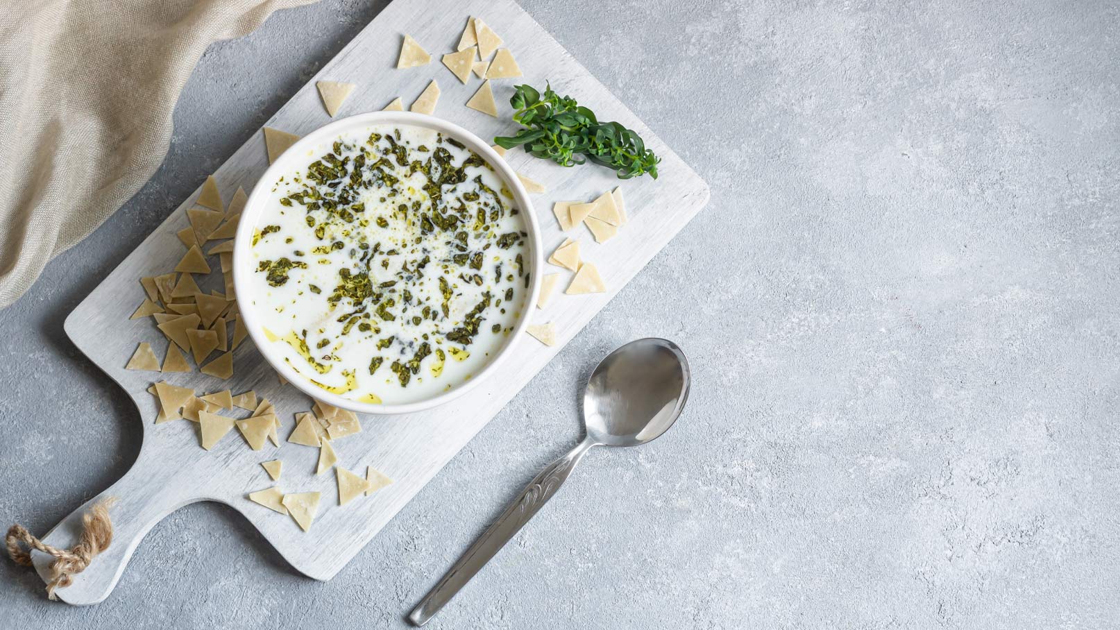 Eine Schüssel mit einer cremigen Suppe, garniert mit grünen Kräutern, steht auf einem Holzbrett. Rund um die Schüssel sind kleine Teigstücke angeordnet, und ein silberner Löffel liegt daneben. Der Hintergrund ist ein neutraler Grauton.