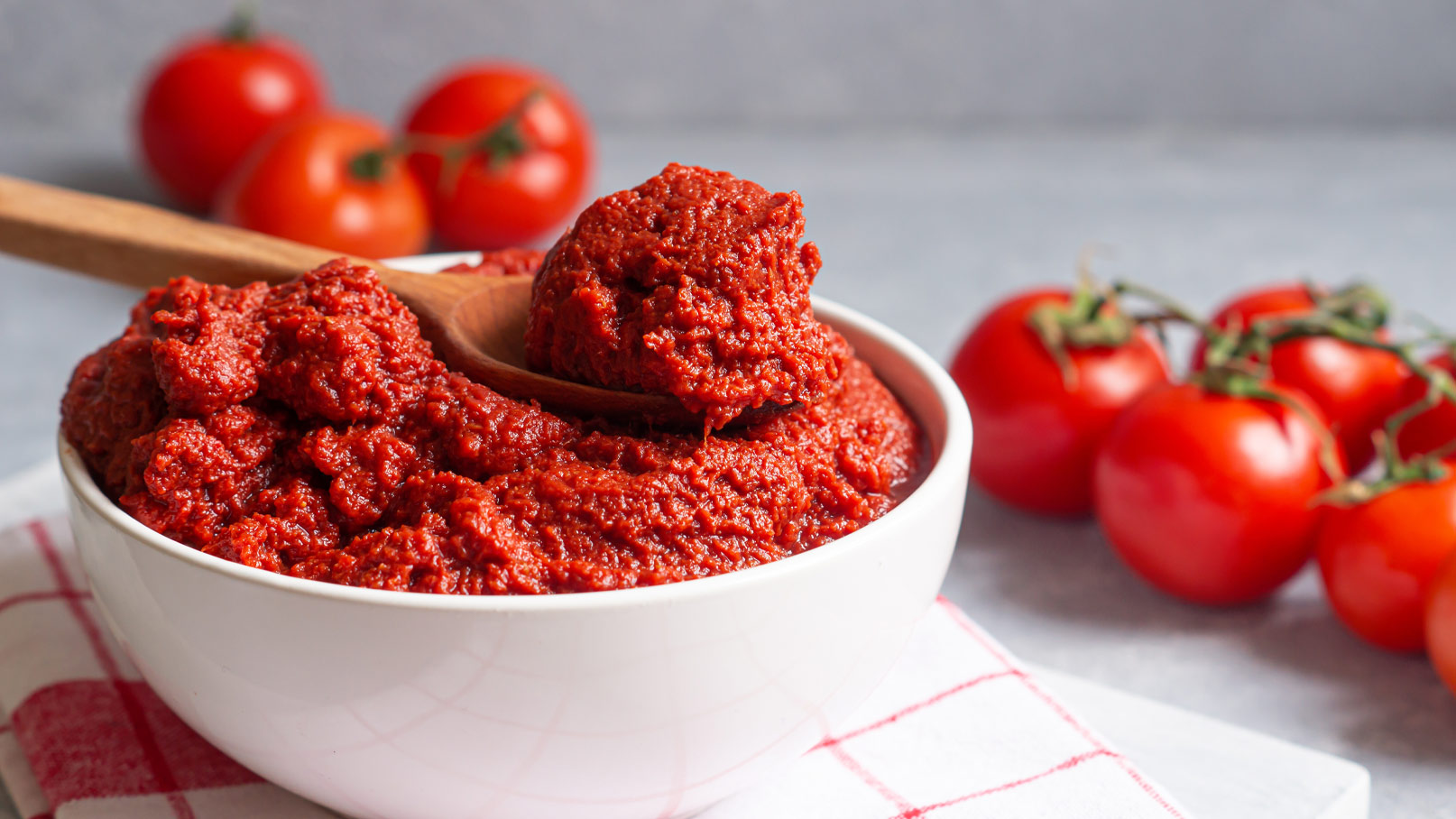 In einer weißen Schüssel befindet sich ein dickes, rotes Tomatenpüree, aus dem ein Löffel herausragt. Im Hintergrund liegen frische, rote Tomaten. Die Szene vermittelt eine einladende Atmosphäre für das Zubereiten von Speisen.