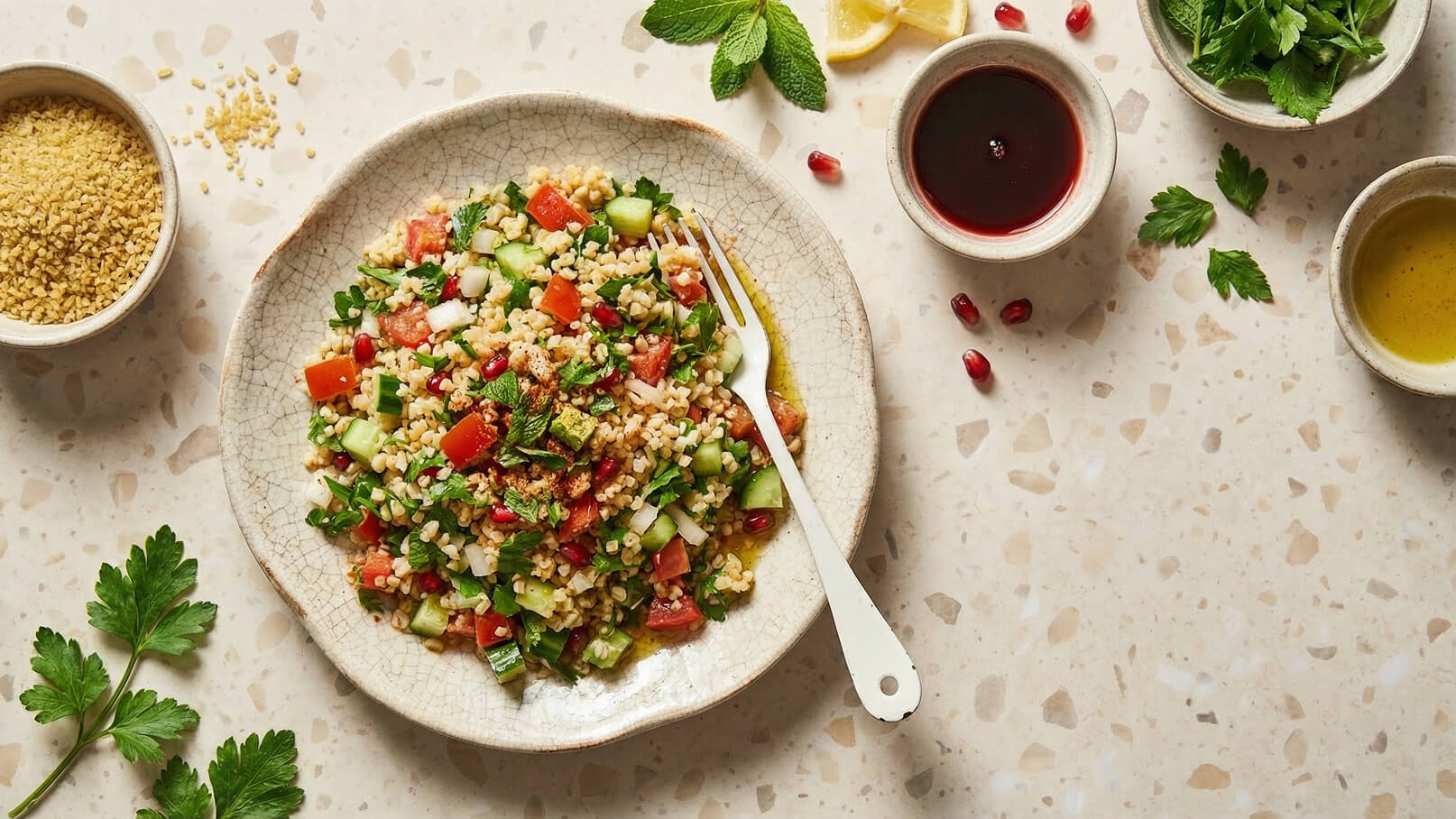 Ein Teller mit frischem Bulgur-Salat, garniert mit Tomaten, Gurken, roten Granatapfelkernen und frischen Kräutern. Eine Gabel steckt im Salat. Neben dem Teller sind kleine Schalen mit Olivenöl und einer dunklen Flüssigkeit sowie eine Schale mit Bulgur zu sehen.
