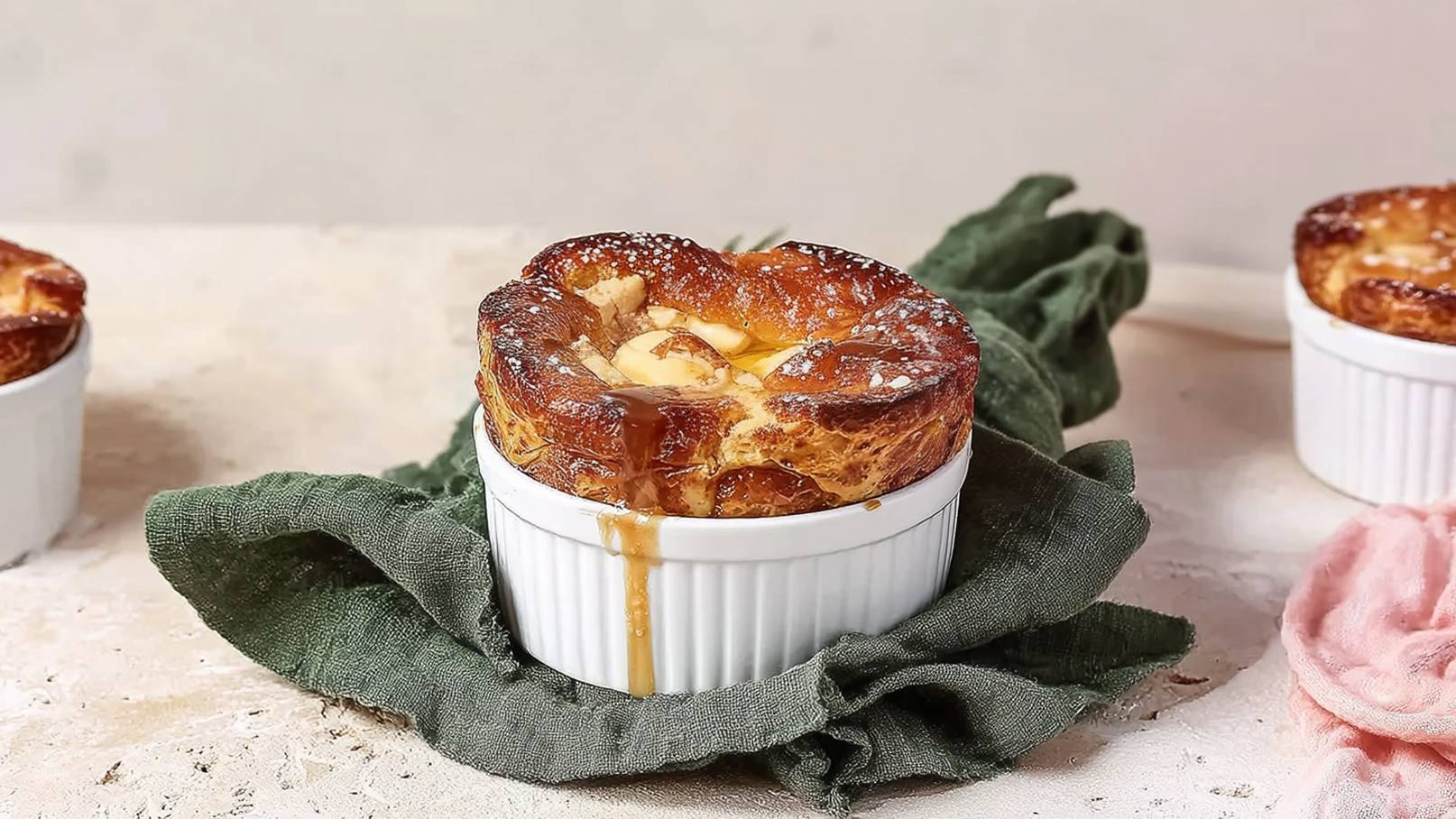 Ein frisch gebackenes Soufflé in einer weißen ramekin. Der oberste Teil ist goldbraun, mit Puderzucker bestäubt. Darunter sieht man eine cremige Füllung, die leicht aus der Form quillt. Das Soufflé liegt auf einem grünen Küchentuch. Im Hintergrund sind weitere ramekins sichtbar.