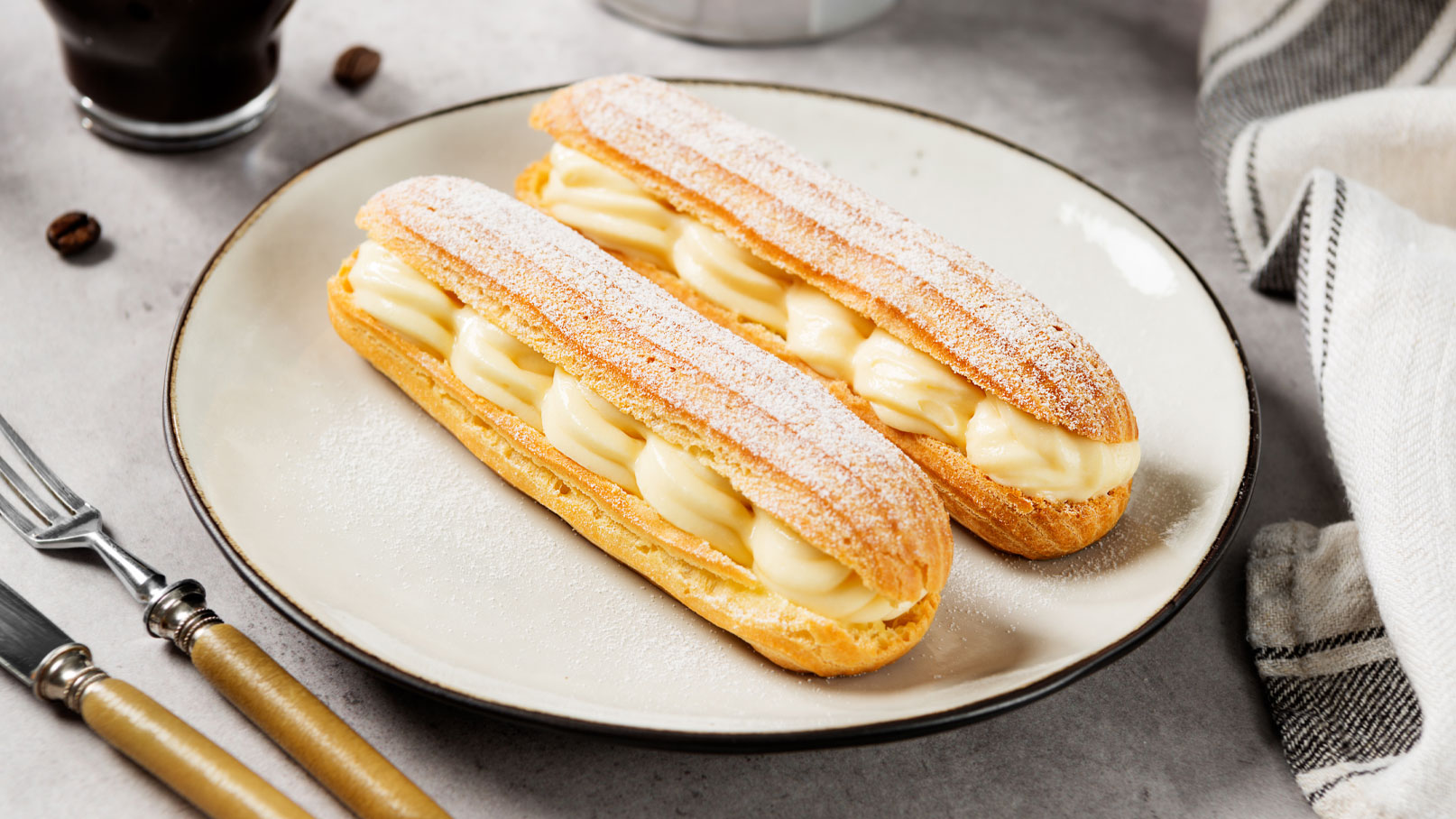 Auf einem weißen Teller liegen zwei Eclairs. Sie sind mit einer glatten, cremigen Füllung gefüllt und mit Puderzucker bestäubt. Neben dem Teller befinden sich Besteck und ein Tuch. Im Hintergrund ist eine Tasse Kaffee zu sehen.
