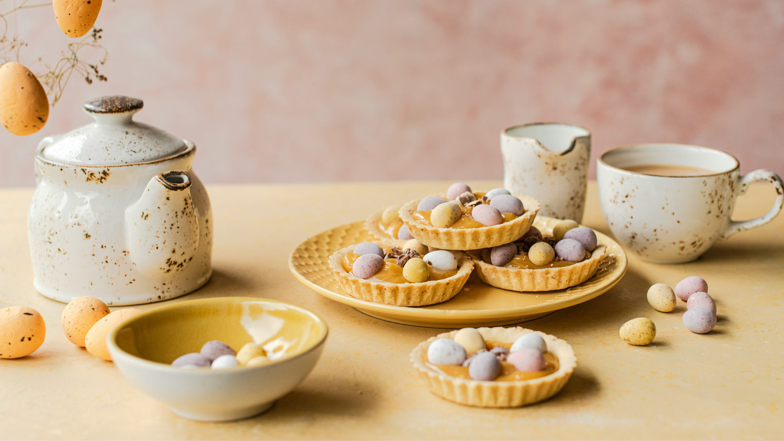 Ein rustikaler Teekessel steht neben einer Tasse Kaffee auf einem Tisch. Darauf sind mehrere knusprige Tartes mit bunten Schokoladeneiern angerichtet, umgeben von einem kleinen Teller mit weiteren Schokoladeneiern und einem erhöhten, gelben Dessertteller.