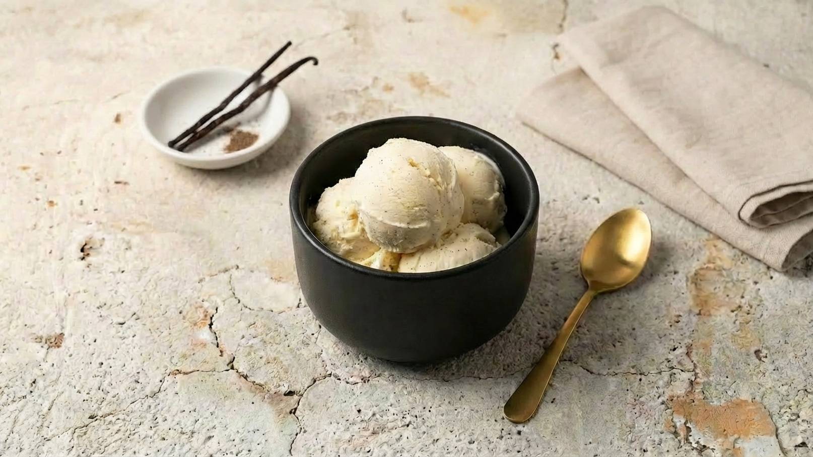 Eine schwarze Schüssel mit mehreren Kugeln Vanilleeis steht auf einer neutralen Oberfläche. Daneben liegt ein kleiner Teller mit Vanilleschoten und ein goldenes Besteckstück. Im Hintergrund ist ein leicht gefaltetes Tuch zu sehen.