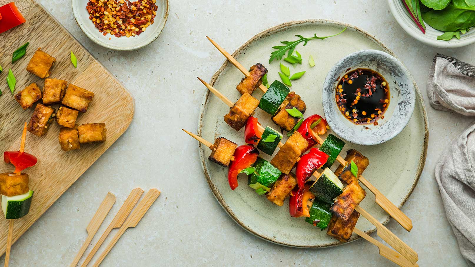 Ein Teller mit gegrillten Spießen aus Tofu, Paprika und Zucchini, serviert mit einer dunklen Soße in einer kleinen Schüssel. Neben dem Teller liegen einige ungenutzte Spieße und ein Holzbrett mit klein geschnittenem Tofu.