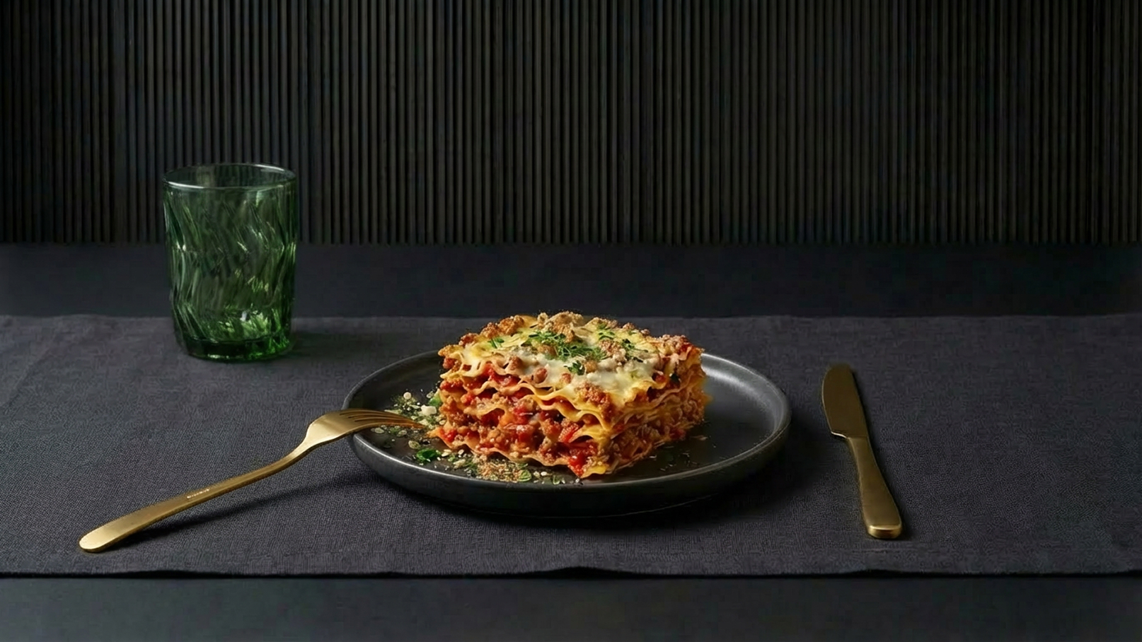 Ein schwarzes Teller mit einer reichhaltigen Lasagne, die Schichten von Nudel, Fleischsoße und geschmolzenem Käse zeigt, steht auf einem dunkelgrauen Tischläufer. Neben dem Teller liegt ein goldener Gabel, und ein grünes Glas steht im Hintergrund.
