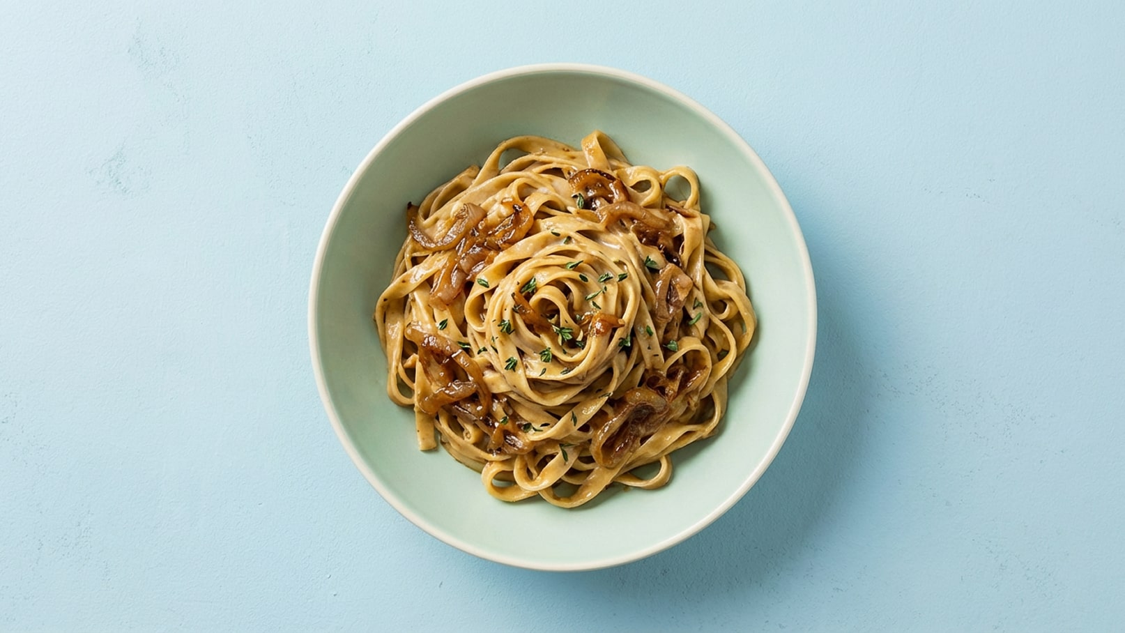 Eine Schüssel mit fettigen, gebratenen Nudeln ist auf einem hellblauen Hintergrund zu sehen, garniert mit karamellisierten Zwiebeln und frischen Kräutern. Die Nudeln sind spiralförmig angerichtet und wirken einladend.
