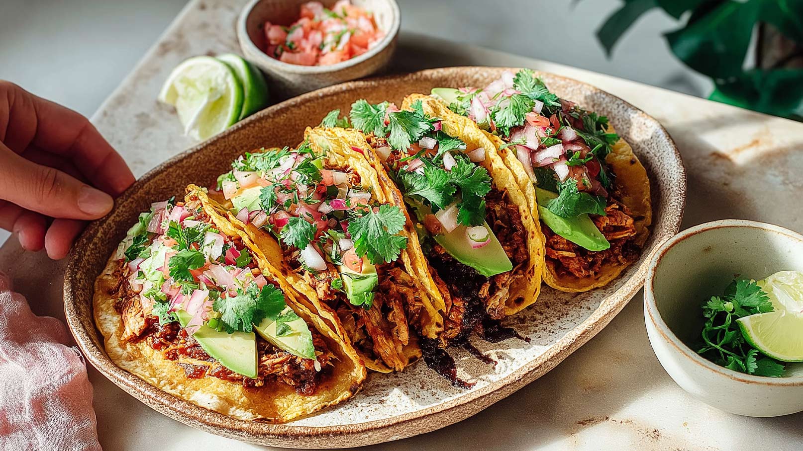 Ein Teller mit fünf Tacos, gefüllt mit zerkleinertem Fleisch und garniert mit frischem Koriander, Zwiebeln und Avocado. Im Hintergrund ist eine kleine Schüssel mit gehackten Tomaten und Limettenscheiben zu sehen. Ein Hand greift nach einem Taco.