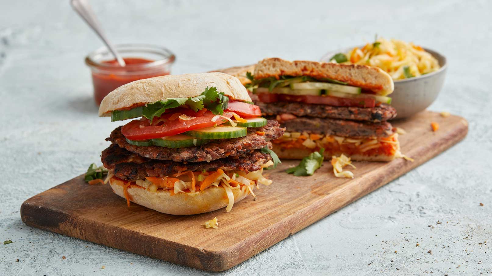 Ein Holzbrett mit zwei üppigen Burgern, gefüllt mit Gemüse wie Tomaten und Gurken. Daneben ist eine Schüssel mit Salat und ein kleines Gefäß mit Soße. Der erste Burger ist aufgeschnitten und zeigt die Schichten im Inneren.