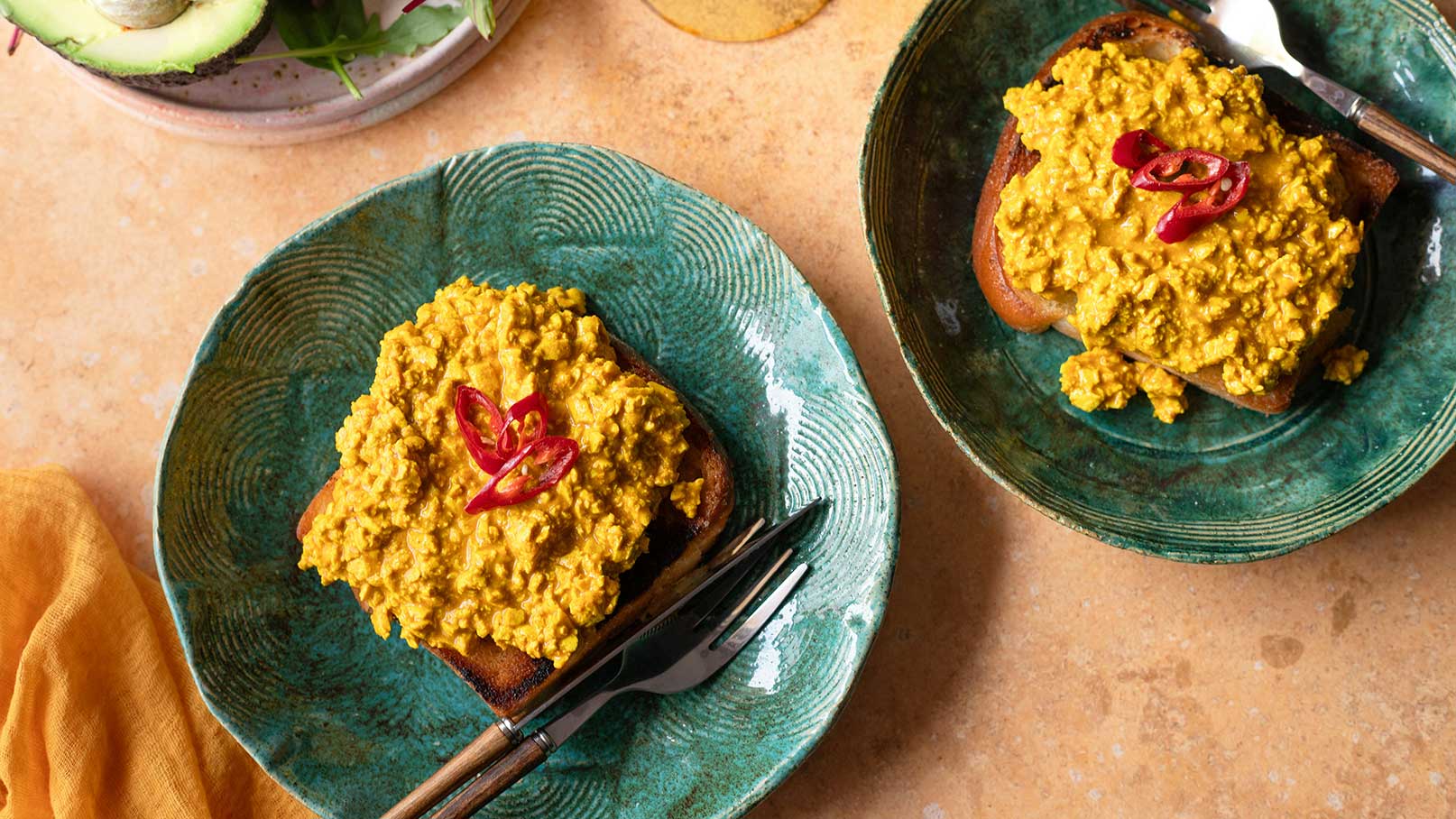 Zwei Teller mit geröstetem Brot, belegt mit einer gelben, cremigen Mischung und rotem Chili als Garnitur. Das Brot auf einem Teller hat eine goldbraune Kruste, während der andere Teller eine ähnliche Belegung zeigt. Hintergrund ist eine sandfarbene Oberfläche.