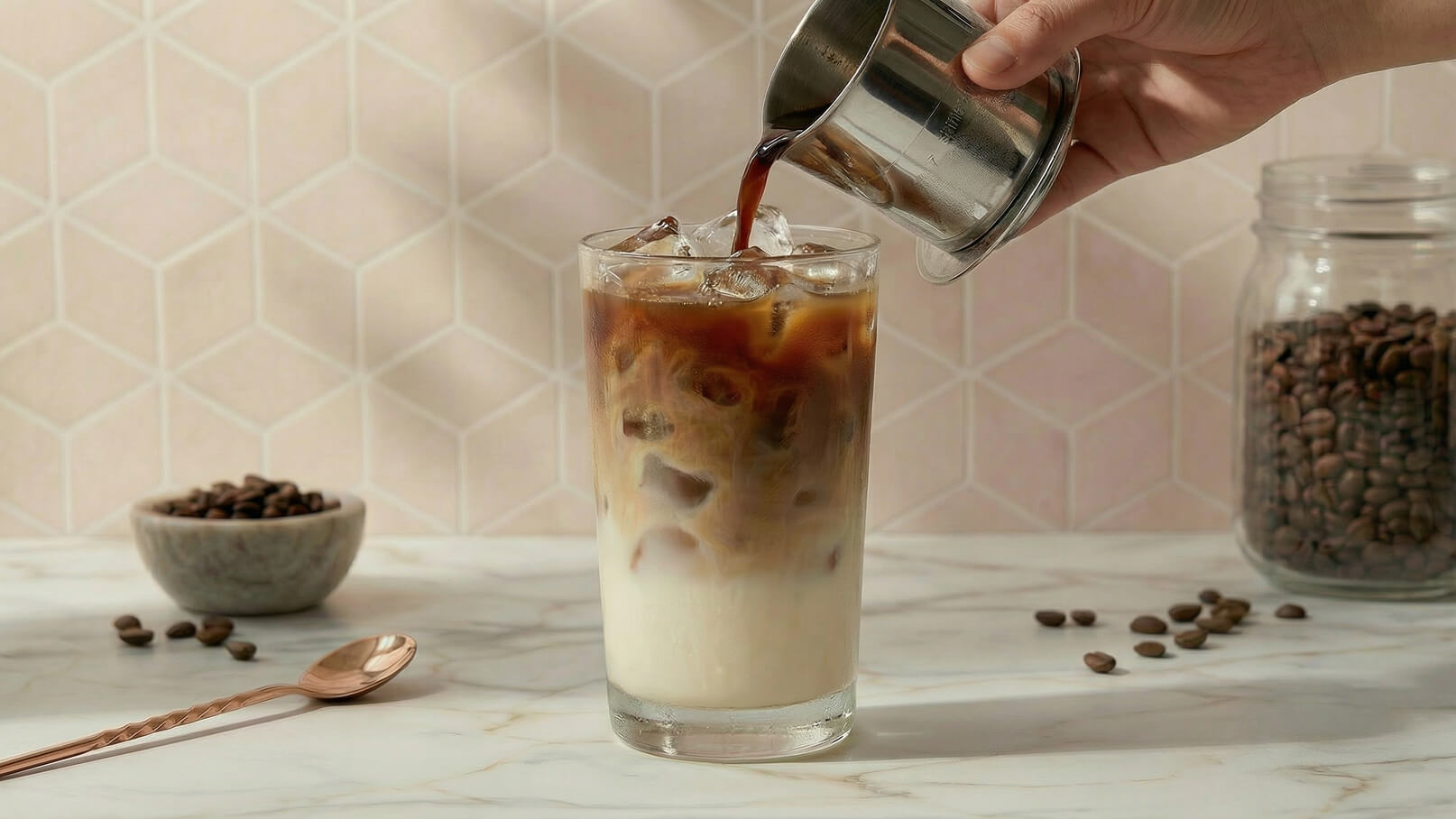 Ein Handheim gießt frischen Kaffee in ein Glas mit Eiswürfeln und Milch. Der Hintergrund zeigt eine helle, geometrische Wand. Eine Schale mit Kaffeebohnen ist neben dem Glas platziert.
