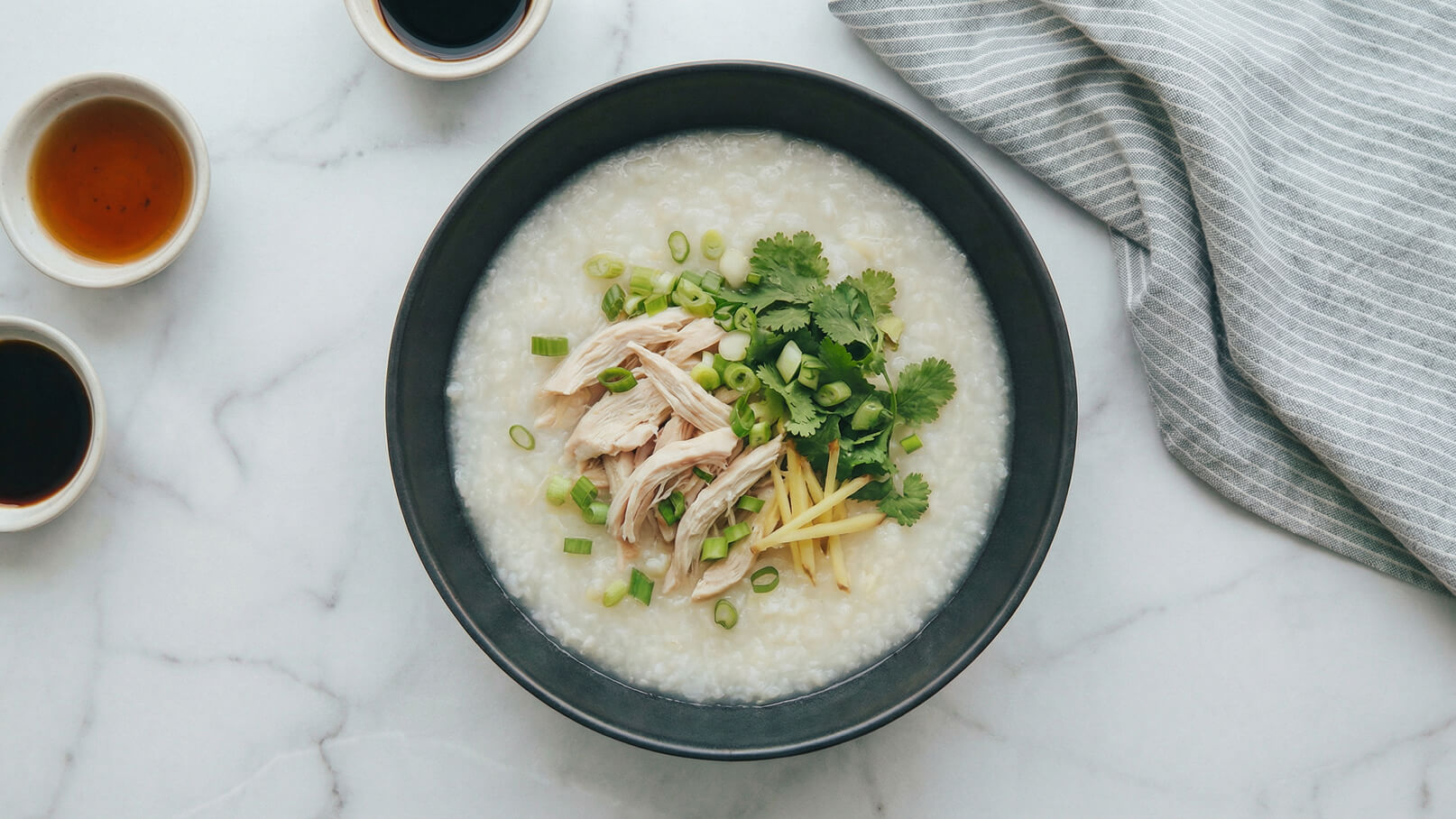 Eine schwarze Schüssel mit Reisbrei, garniert mit zarten Hähnchenstücken, frischen Korianderblättern, geschnittenem Ingwer und Frühlingszwiebeln. Im Hintergrund befinden sich kleine Schalen mit verschiedenen Saucen. Der Tisch hat eine helle Marmoroberfläche.