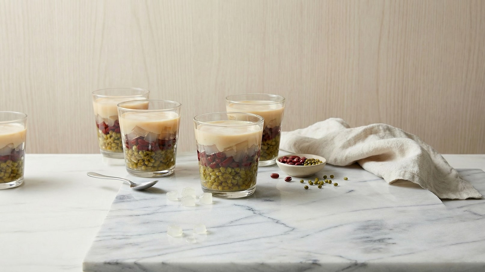 Fünf Gläser mit geschichteten Zutaten stehen auf einem marmorierten Tisch. Die Gläser enthalten grüne und rote Bohnen, transparente Gelee-Stücke und eine cremige Schicht darüber. Neben den Gläsern liegen ein Löffel und einige Bohnen verstreut auf dem Tisch. Ein Tuch ist leicht zusammengelegt.