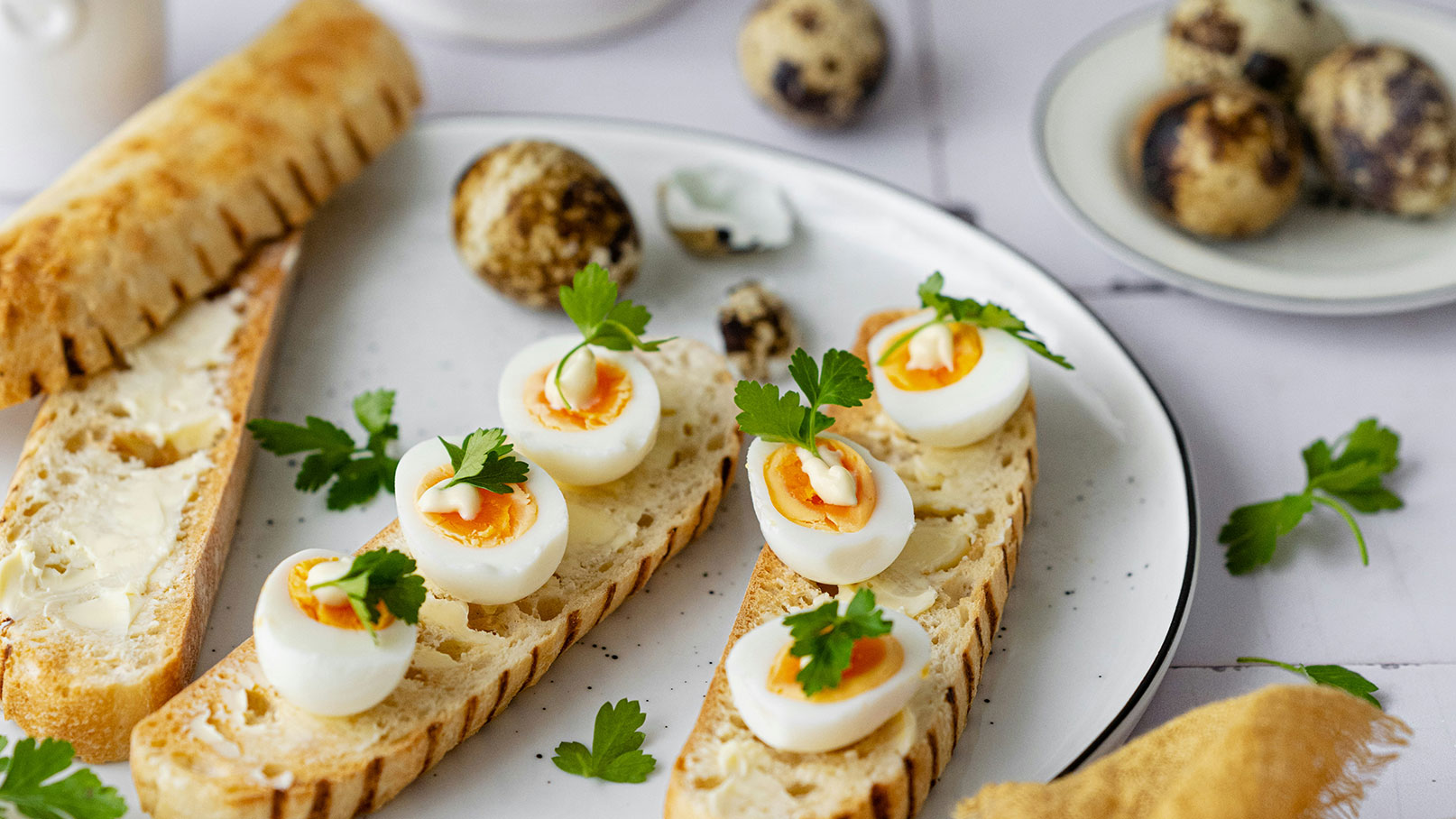 Ein Teller mit geröstetem Brot, belegt mit kleinen, halben Eiern und frischer Petersilie. Die Eier haben eine orangefarbene Füllung und sind mit einer weißen Soße verziert. Neben dem Teller liegen mehrere Wachtel-Eier.