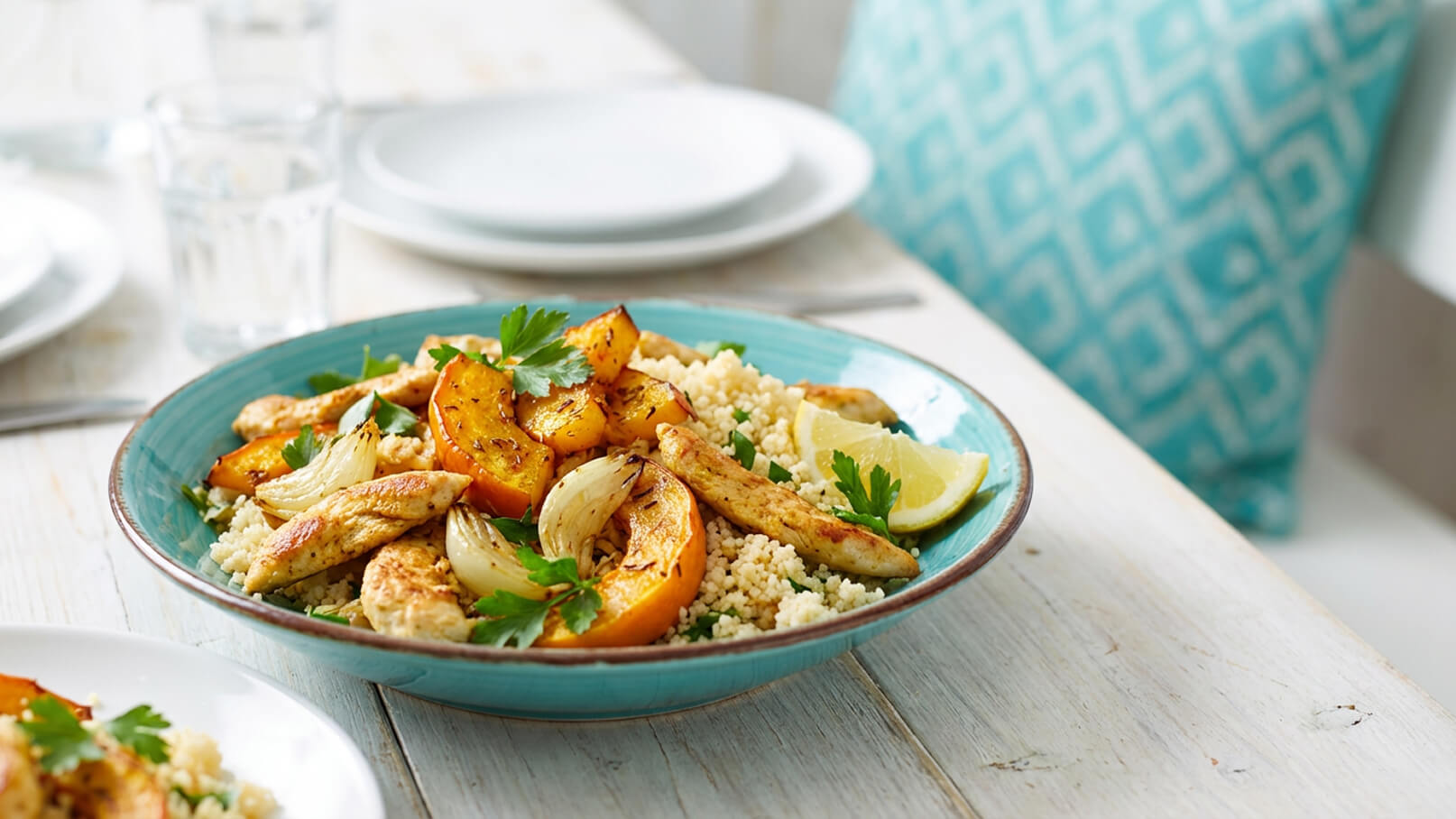 Ein Teller mit Couscous, garniert mit gebratenem Hähnchen, gegrilltem Kürbis und Zwiebeln. Frische Petersilie und Zitronenscheiben verleihen dem Gericht zusätzlichen Geschmack. Der Teller hat eine türkisfarbene Umrandung.