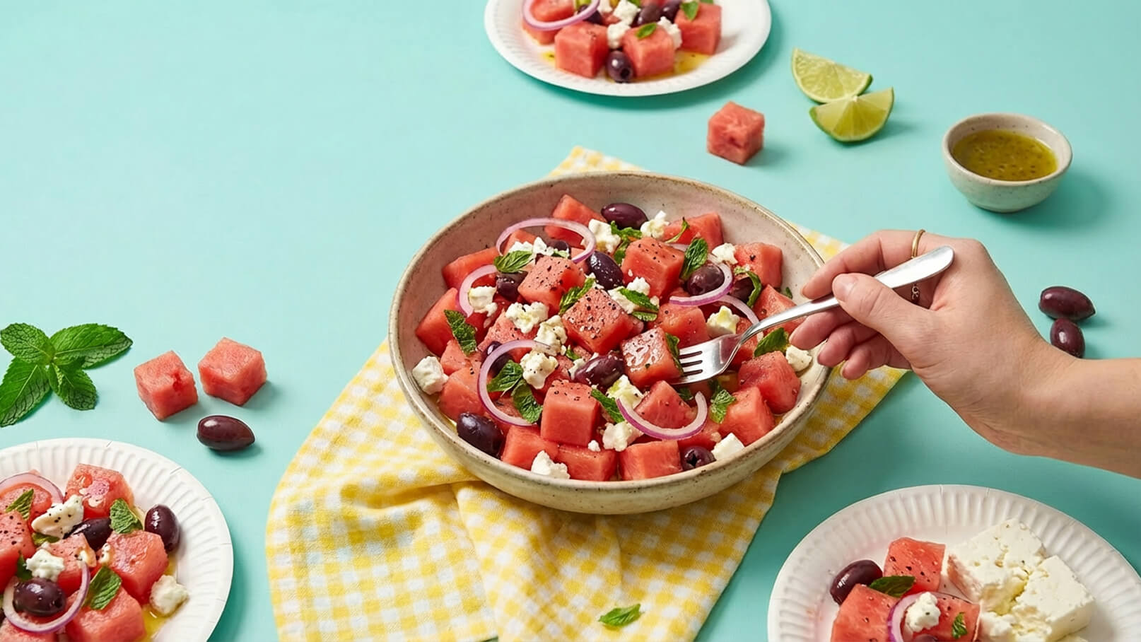 Eine Schüssel mit Wassermelonensalat, garniert mit Oliven, Feta, roten Zwiebeln und frischer Minze. Eine Hand mit einer Gabel nimmt einen Bissen aus der Schüssel. Der Salat liegt auf einem karierten Geschirrtuch, umgeben von weiteren Tellern und Limettenscheiben.