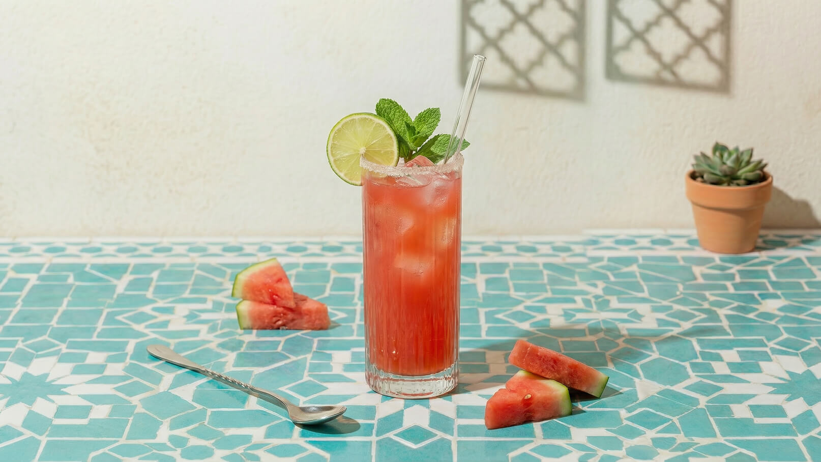 Ein hoher Cocktail mit Wassermelone, Limette und Minze steht auf einem blauen, mit geometrischen Mustern dekorierten Tisch. Neben dem Getränk liegen einige Stücke Wassermelone und ein Löffel.
