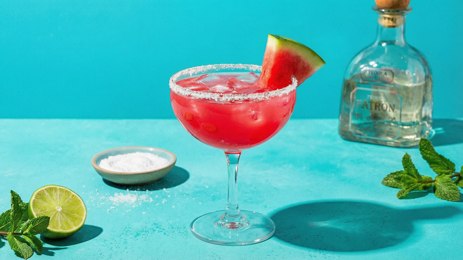 Ein buntes Glas mit einem pinken Cocktail, der mit Eis gefüllt ist und an den Rändern mit Salz bestreut ist. Ein Stück Wassermelone dekoriert das Glas. Neben dem Cocktail sind eine halbe Limette und eine Schale mit weißem Salz. Der Hintergrund ist in kräftigem Türkis gehalten.