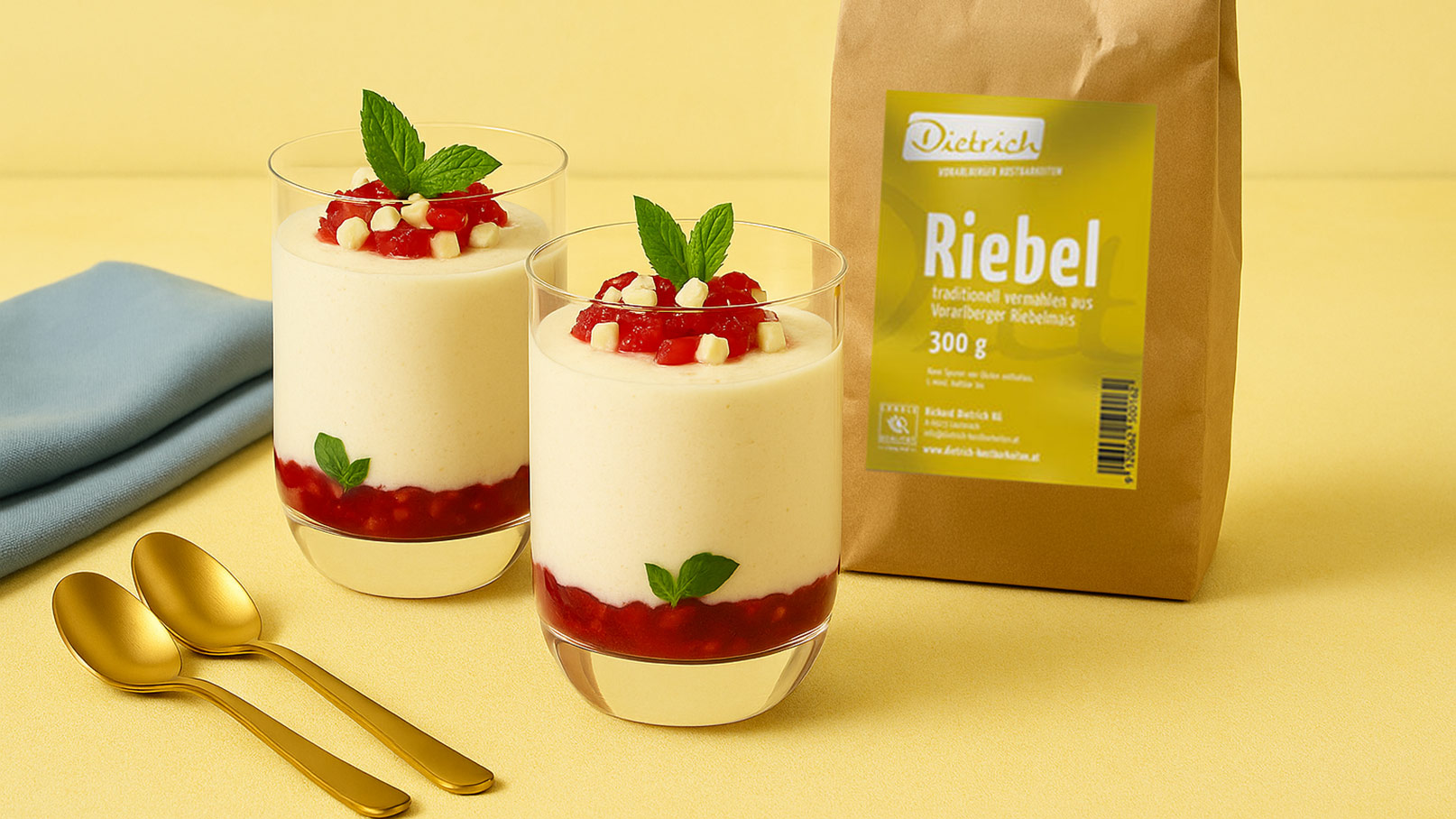 Zwei Gläser mit einer schaumigen Dessertcreme, verziert mit roter Fruchtsoße und Minzblättern. Neben den Gläsern liegen zwei goldene Löffel und eine Packung mit der Aufschrift "Riebel" auf einem gelben Untergrund.