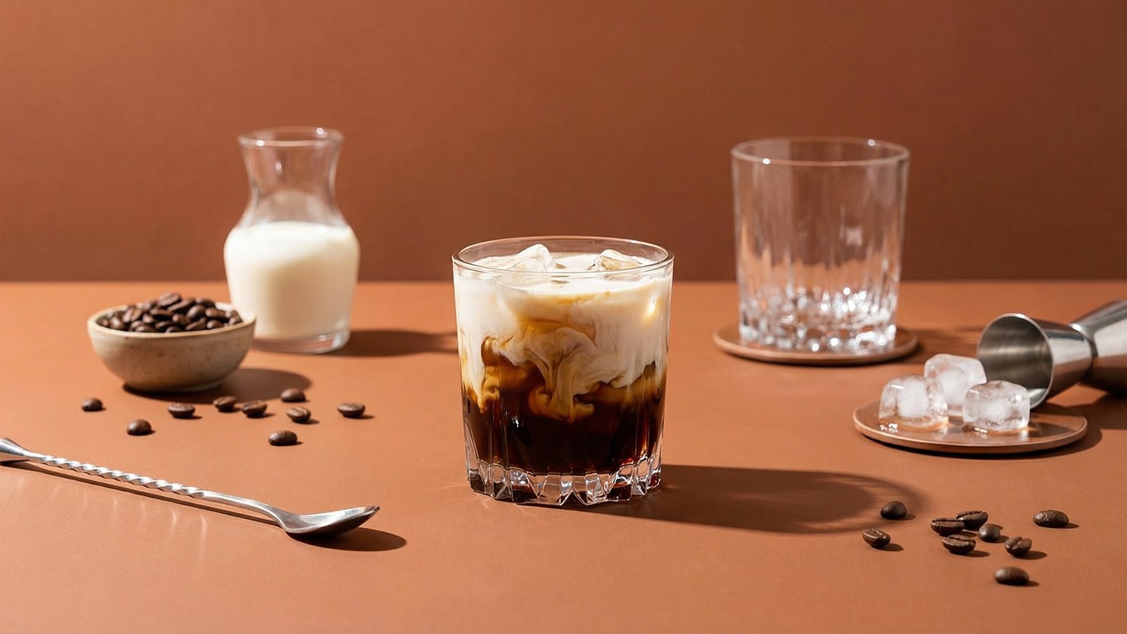Ein Glas mit einem Eiskaffee, der aus dunkler Flüssigkeit und milchiger Schichtung besteht, steht im Vordergrund. Im Hintergrund sind eine Milchkanne, eine Schale mit Kaffeebohnen und ein weiteres Glas zu sehen. Der Tisch hat eine braune Tischoberfläche.