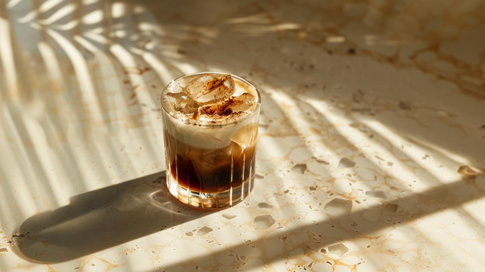 Ein Glas mit einem bunten Cocktail steht auf einer Theke. Der Drink hat eine schaumige Oberfläche, Eiswürfel und ist mit einer Kirsche und einer Zitronenscheibe garniert. Im Hintergrund sind verschwommene Flaschen zu erkennen.
