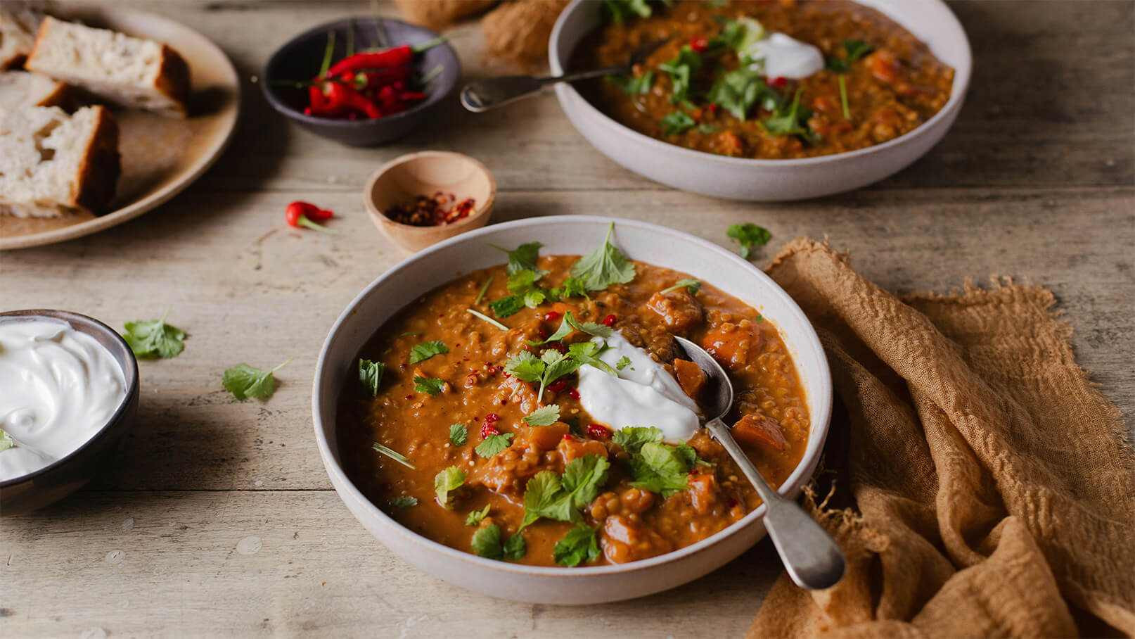 Zwei Schalen mit dampfender Suppe, garniert mit frischem Koriander und einem Klecks Joghurt, stehen auf einem Holztisch. Daneben sind Schalen mit Gewürzen und einige Stücke Brot zu sehen.