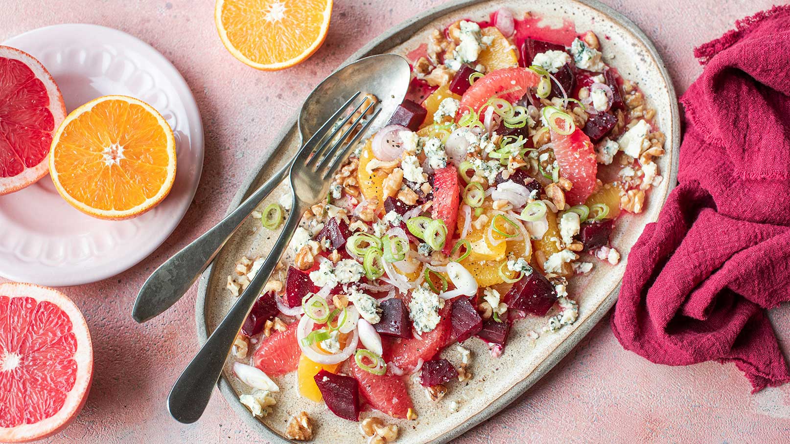 Ein gedeckter Teller mit einem bunten Salat aus verschiedenen Zitrusfrüchten, Rote Bete, Zwiebeln, Walnüssen und zerbröseltem Käse. Zwei Gabeln liegen auf dem Teller, während im Hintergrund halbierte Grapefruits und eine Orange sichtbar sind. Der Untergrund ist rosa.