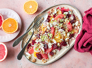 Ein Teller mit buntem Fruchtsalat, der aus verschiedenen Obstsorten wie Grapefruit, Orange und Rote Bete besteht. Darüber sind gehackte Frühlingszwiebeln und zerbröselter Käse verteilt. Daneben liegen frische, halbierte Orangen und eine rote Serviette. Der Hintergrund ist hell und farbenfroh.