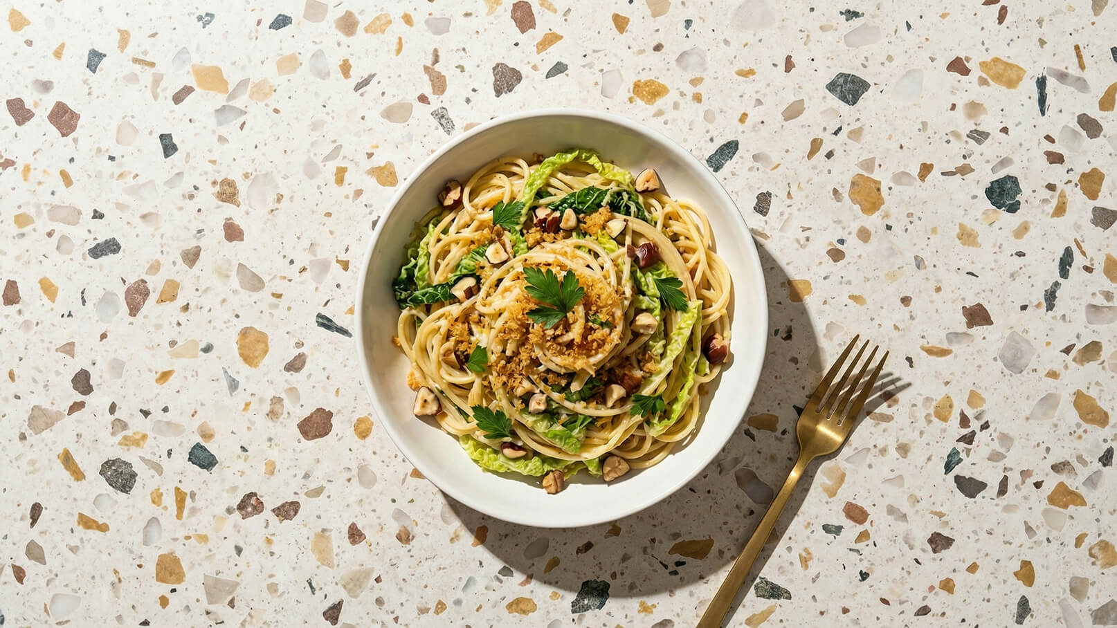 Ein Teller mit Spaghetti, garniert mit gehacktem Gemüse, Nüssen und frischer Petersilie. Die Pasta liegt auf einem Bett aus grünen Blättern. Der Teller ist vor einem hellen, gemusterten Untergrund platziert. Eine goldene Gabel liegt neben dem Teller.