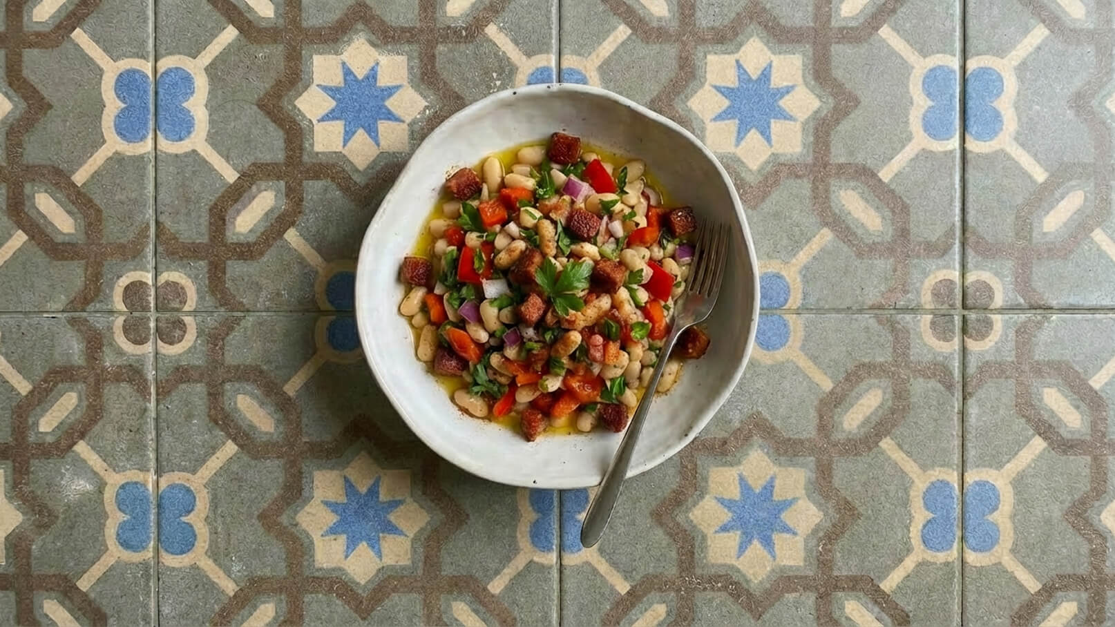 Eine weiße Schüssel mit einem bunten Bohnensalat ist im Bild zu sehen. Der Salat enthält weiße und rote Bohnen, gewürfelte Paprika, Zwiebeln und frische Kräuter. Ein Besteck liegt daneben. Der Hintergrund zeigt ein dekoratives Fliesenmuster.