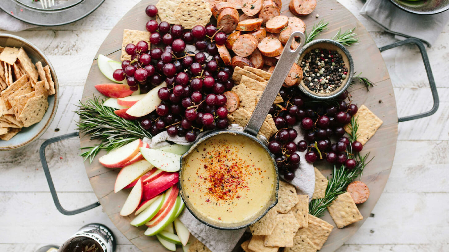 Eine rustikale Platte mit verschiedenen Snacks. Im Mittelpunkt befindet sich eine Schüssel mit cremigem Dip, umgeben von geschnittenen Äpfeln, Trauben, Crackern und Wurstscheiben. Frische Rosmarinzweige schmücken die Anordnung, die einladend und abwechslungsreich wirkt.